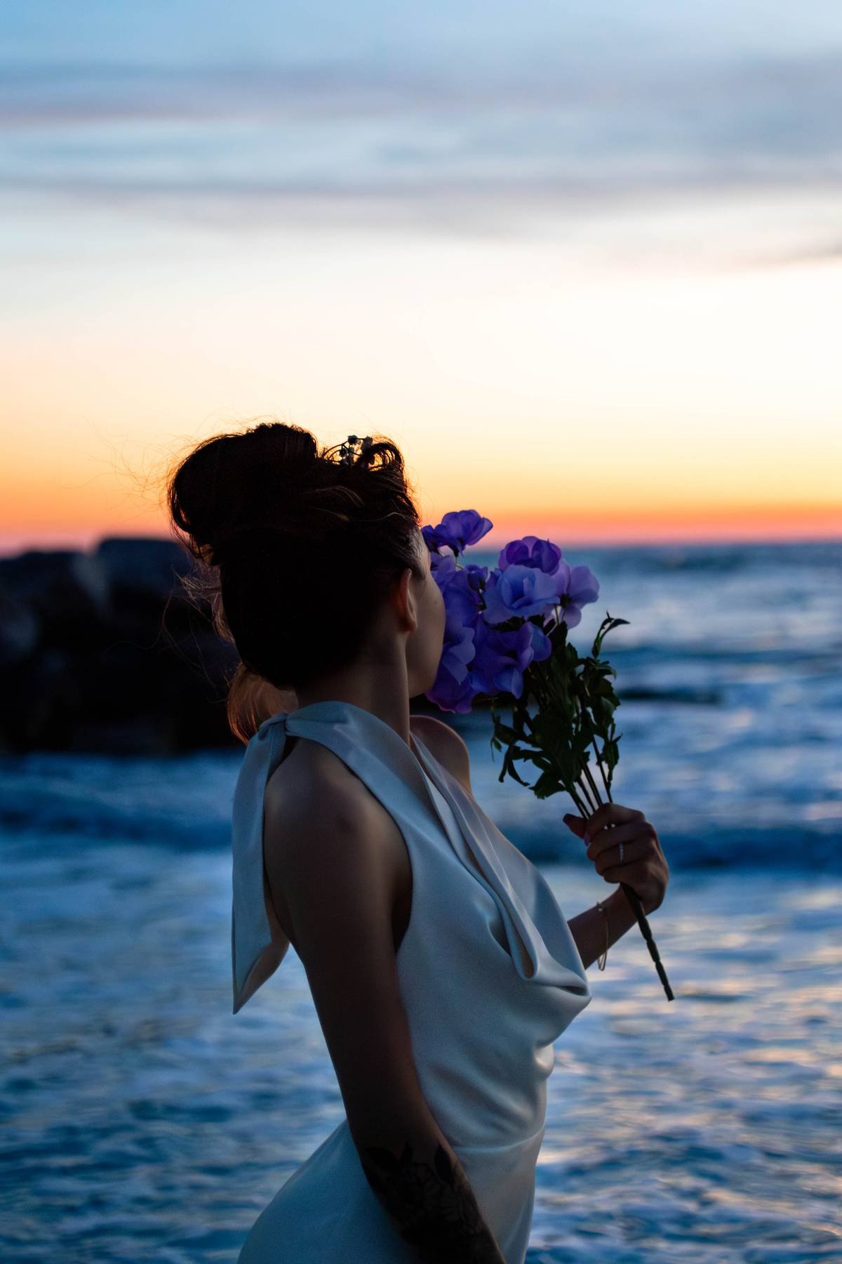 A woman with dark hair styled in an updo holding a bouquet of purple flowers while standing by the ocean during sunset.
