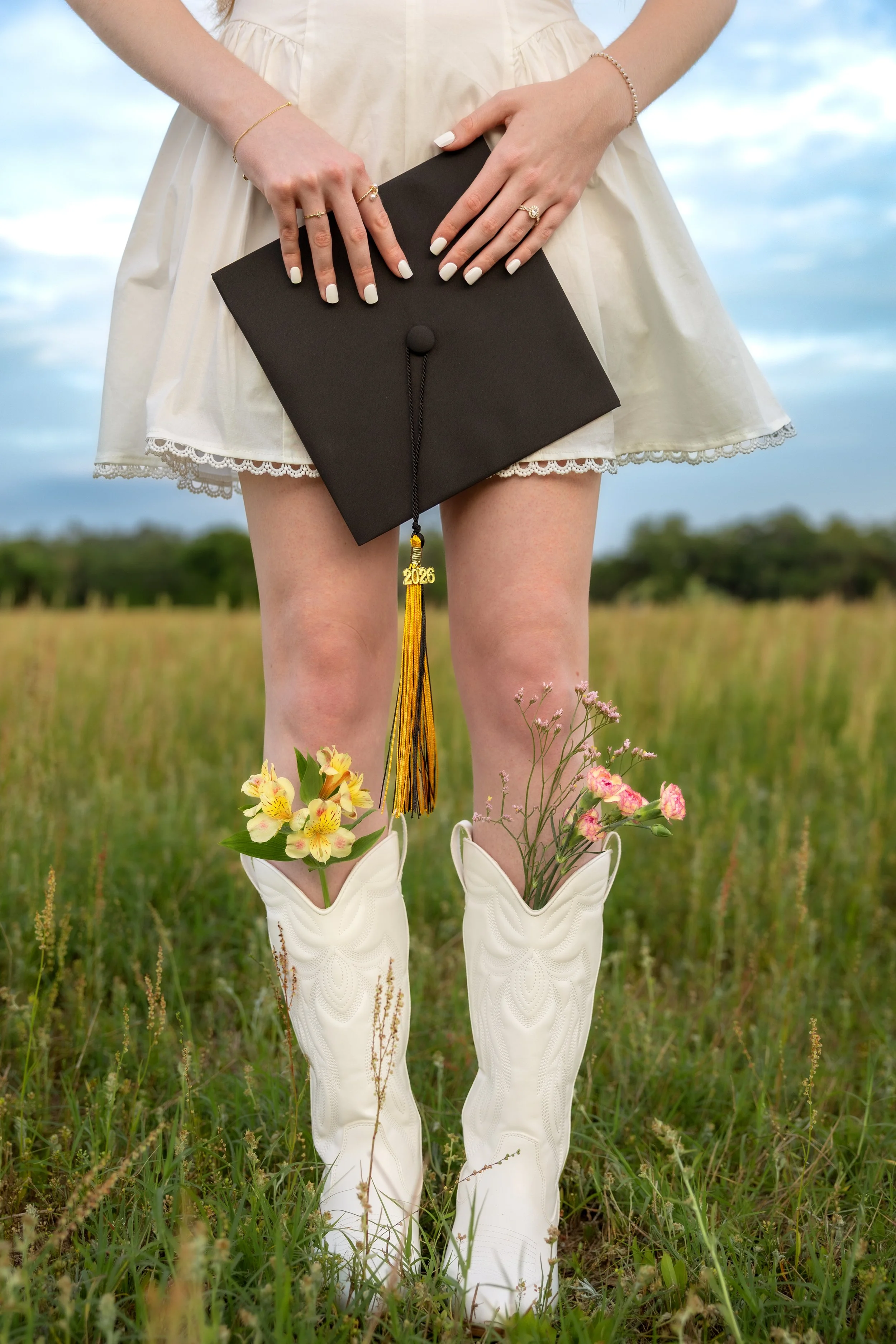 A person dressed in a white dress and cowboy boots standing in a grassy field holding a black graduation cap with a gold tassel that has '2026' attached. The person's hands are visible, adorned with rings and bracelets, with flowers tucked into the b