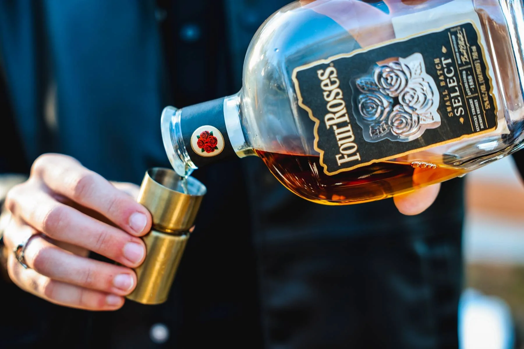 Pouring tequila from a bottle labeled 'Four Roses' into a shot glass held by a person's hand.
