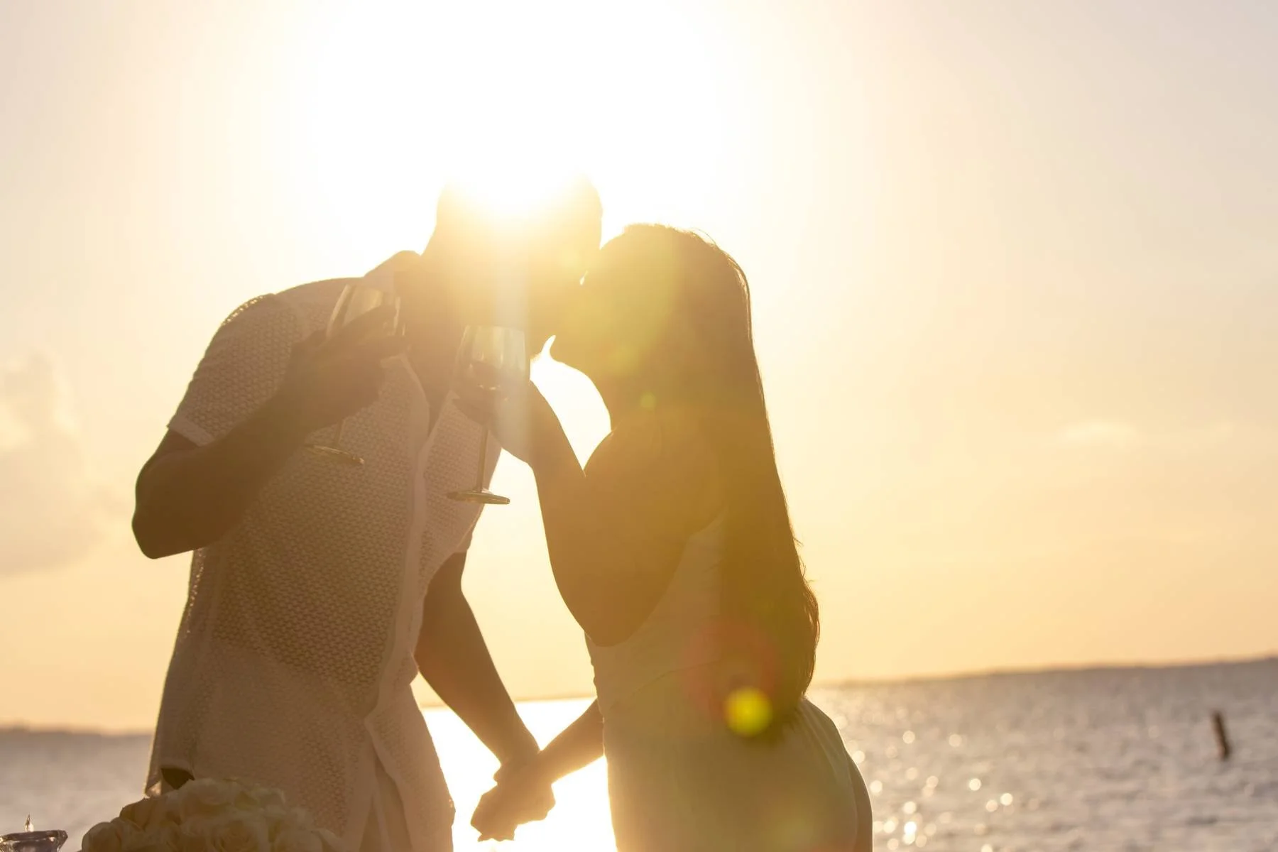 A silhouette of a couple standing outdoors at sunset, kissing and holding hands, with the ocean in the background.