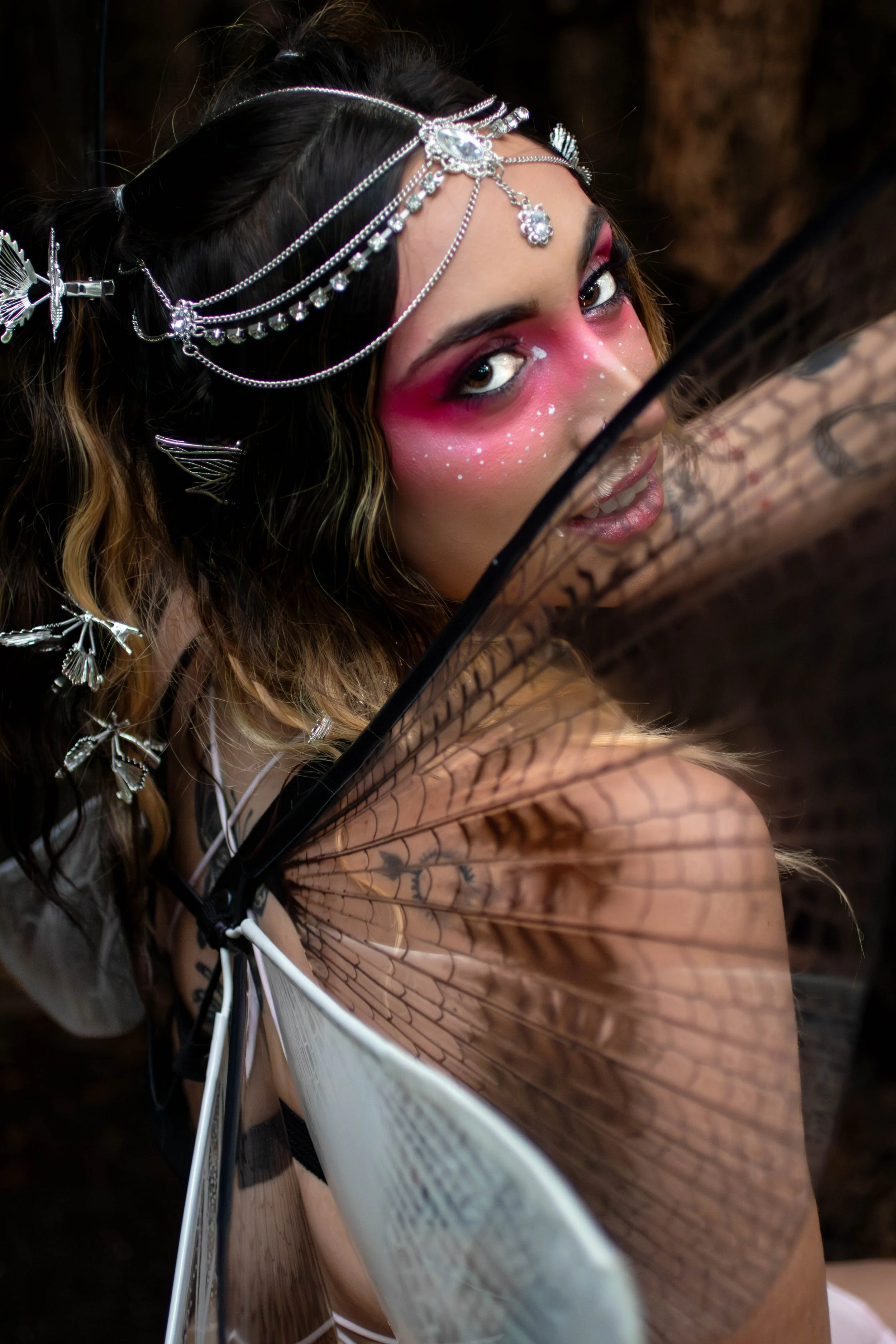 A woman with pink and white face makeup, silver jewelry, and butterfly wings holding a black lace fan.