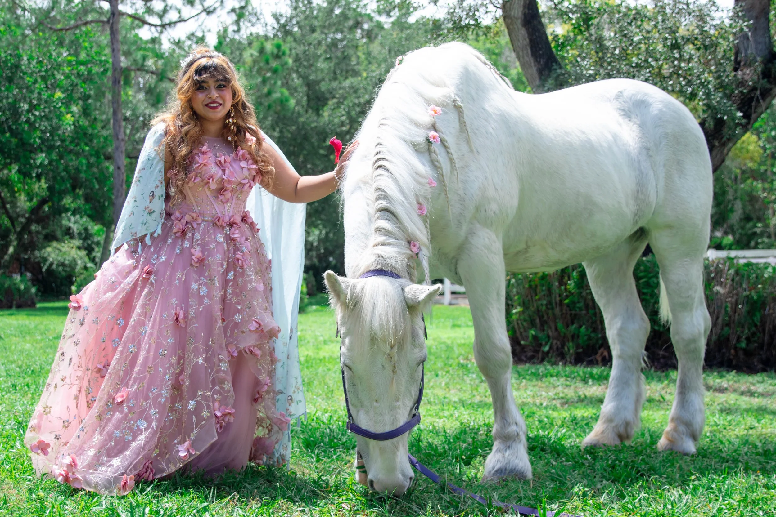 A woman dressed in a pink, floral gown petting a white horse decorated with pink flowers in a lush green outdoor setting.