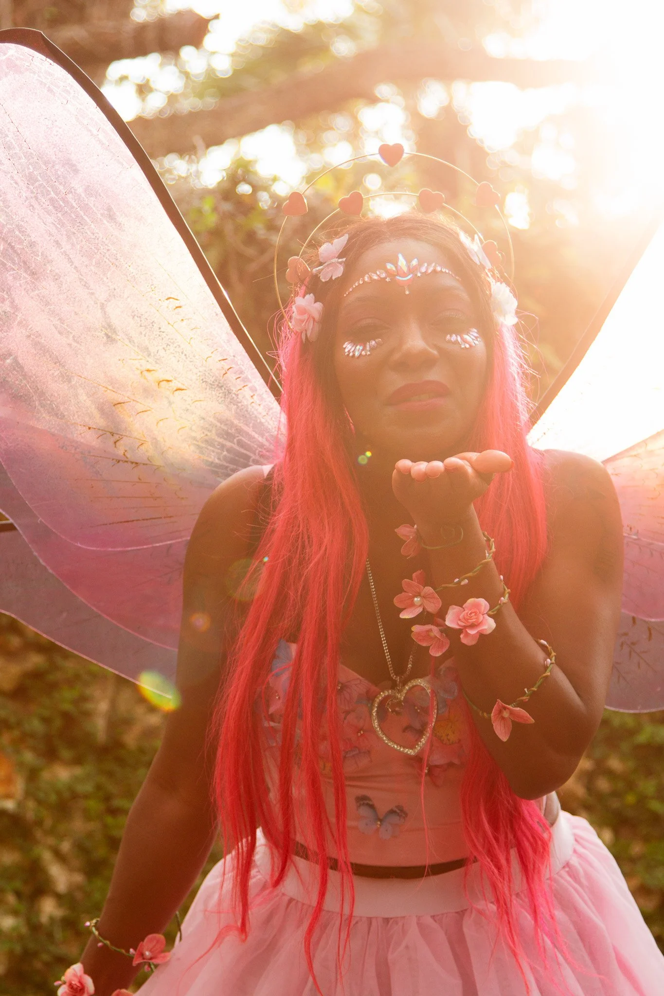 A person dressed as a fairy blowing a kiss in a forest setting with sunlight filtering through the trees. They have long pink hair, floral and glittery makeup, and are wearing fairy wings, a floral crown, and a pink tutu skirt.