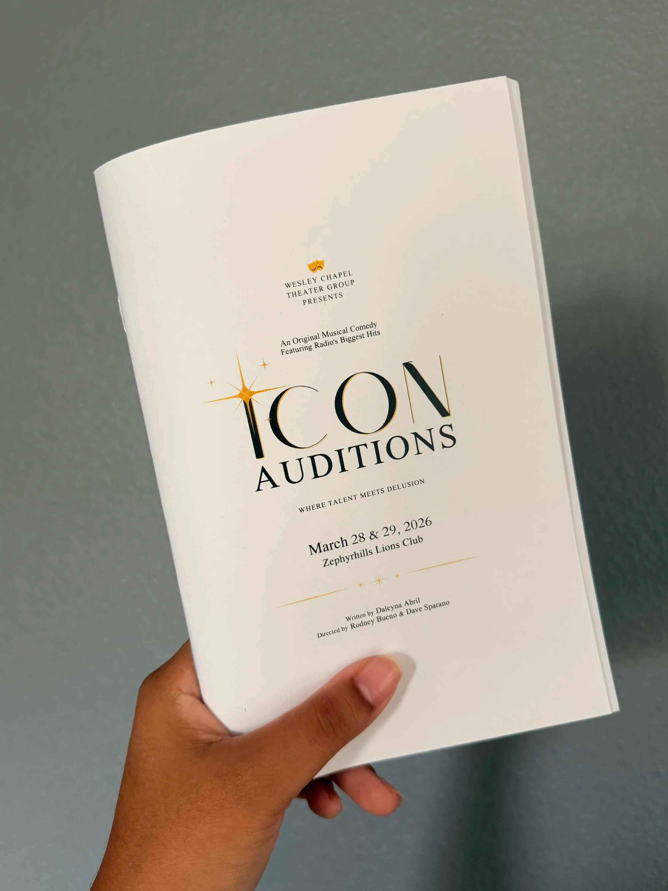 Hand holding a brochure for a musical comedy audition event titled Icons Auditions, scheduled for March 28 and 29, 2026 at Zephyrhills Lions Club, written by Daleyna Abril, directed by Rodney Bueno and Dave Sparano.