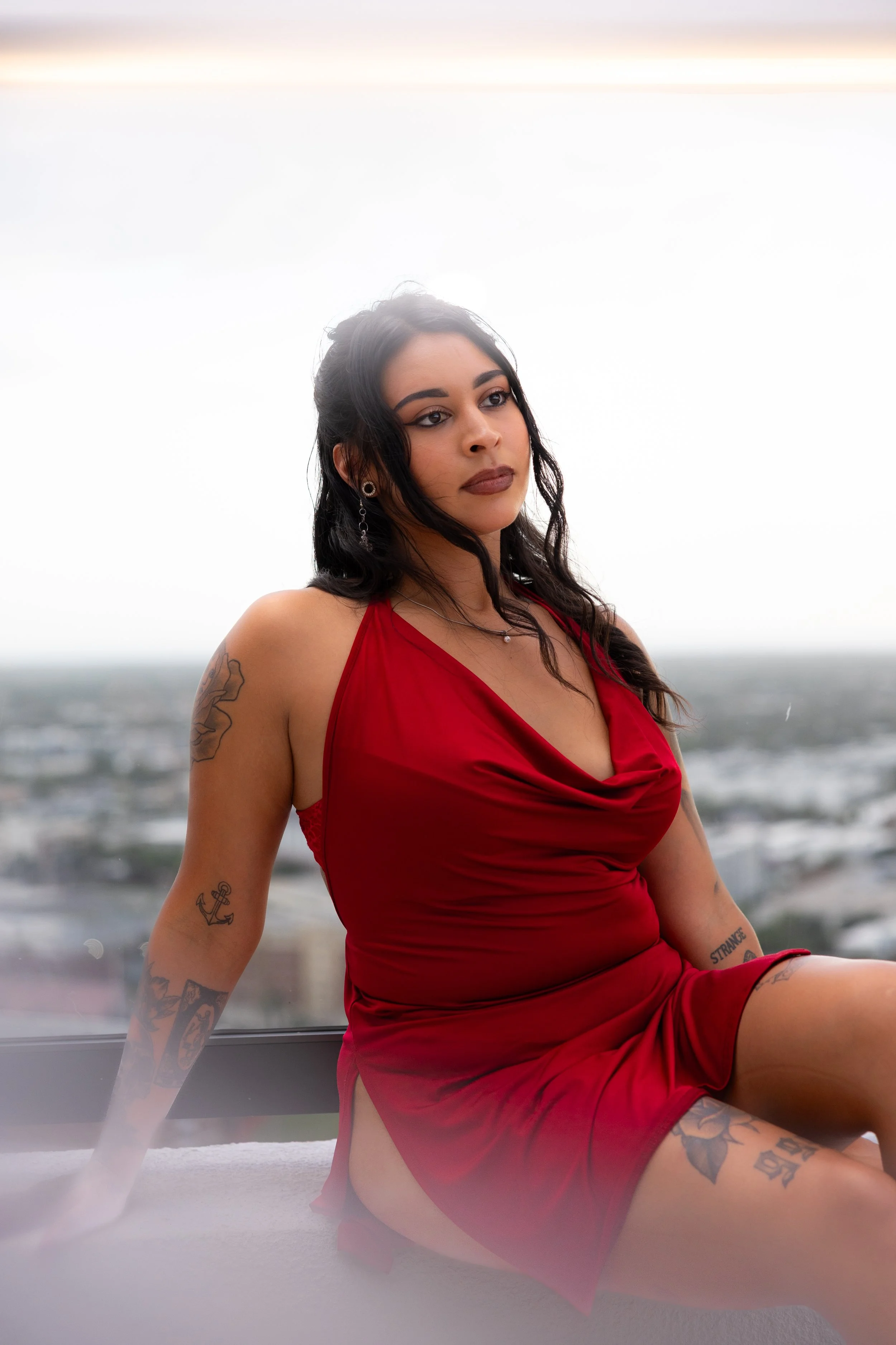 A woman with dark hair and tattoos wearing a red dress, sitting indoors near a large window. The background includes a blurred view of a cloudy sky and cityscape.