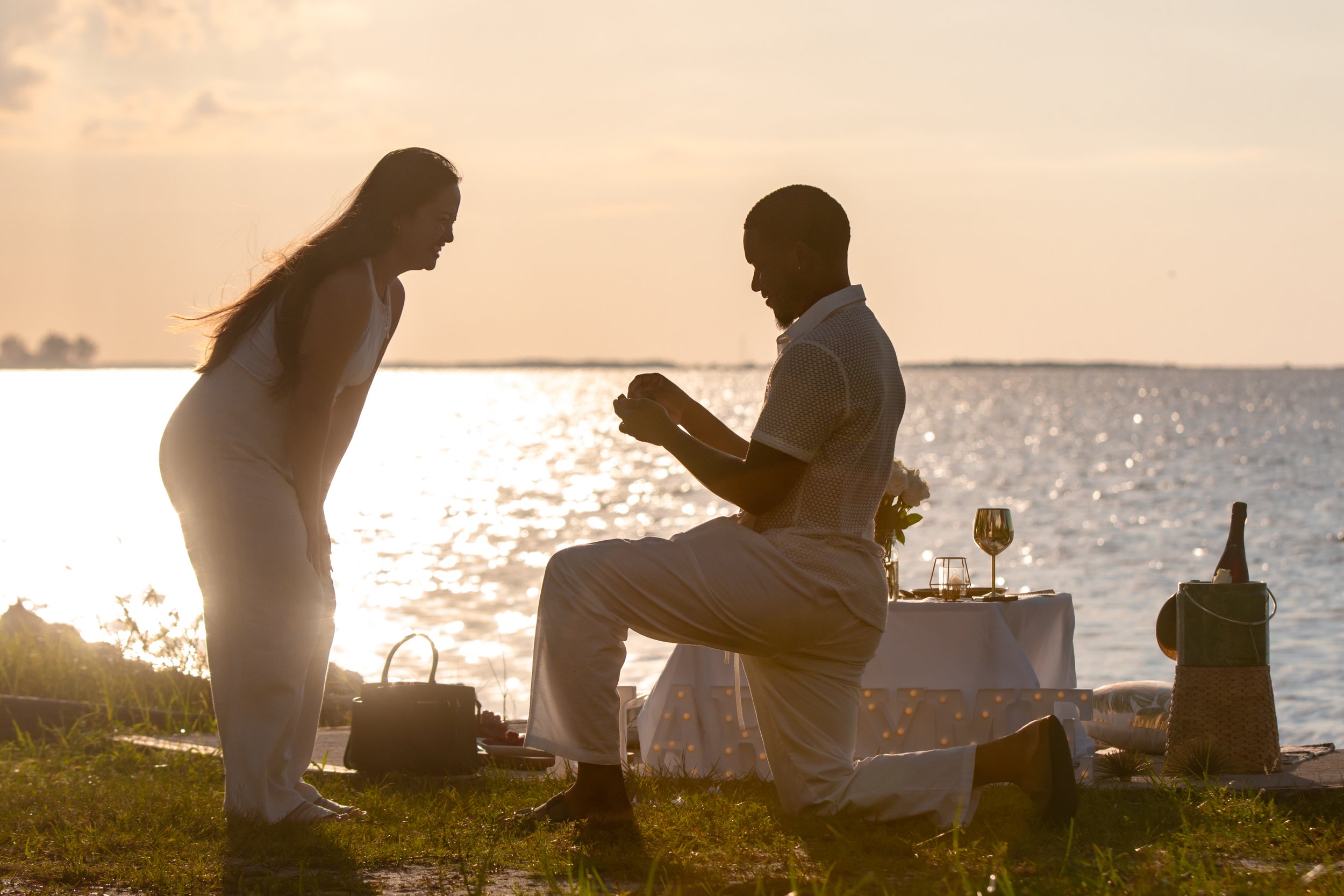A man proposing marriage to a woman at sunset by a body of water, with a table set for a romantic dinner nearby.