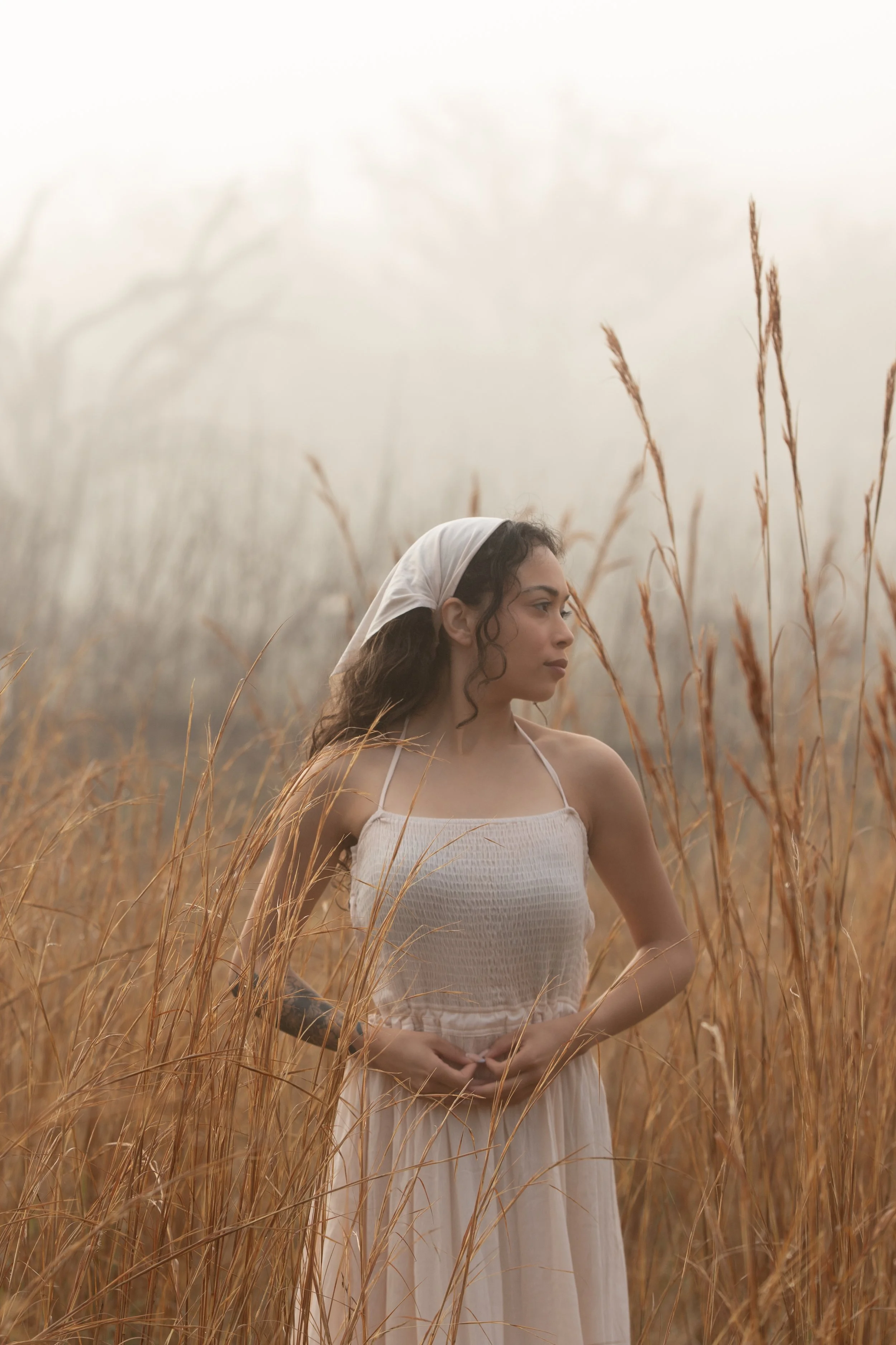 A young woman with dark curly hair wearing a beige sleeveless dress and a white headscarf stands in a field of tall, dry grass, looking to the right with a calm expression.