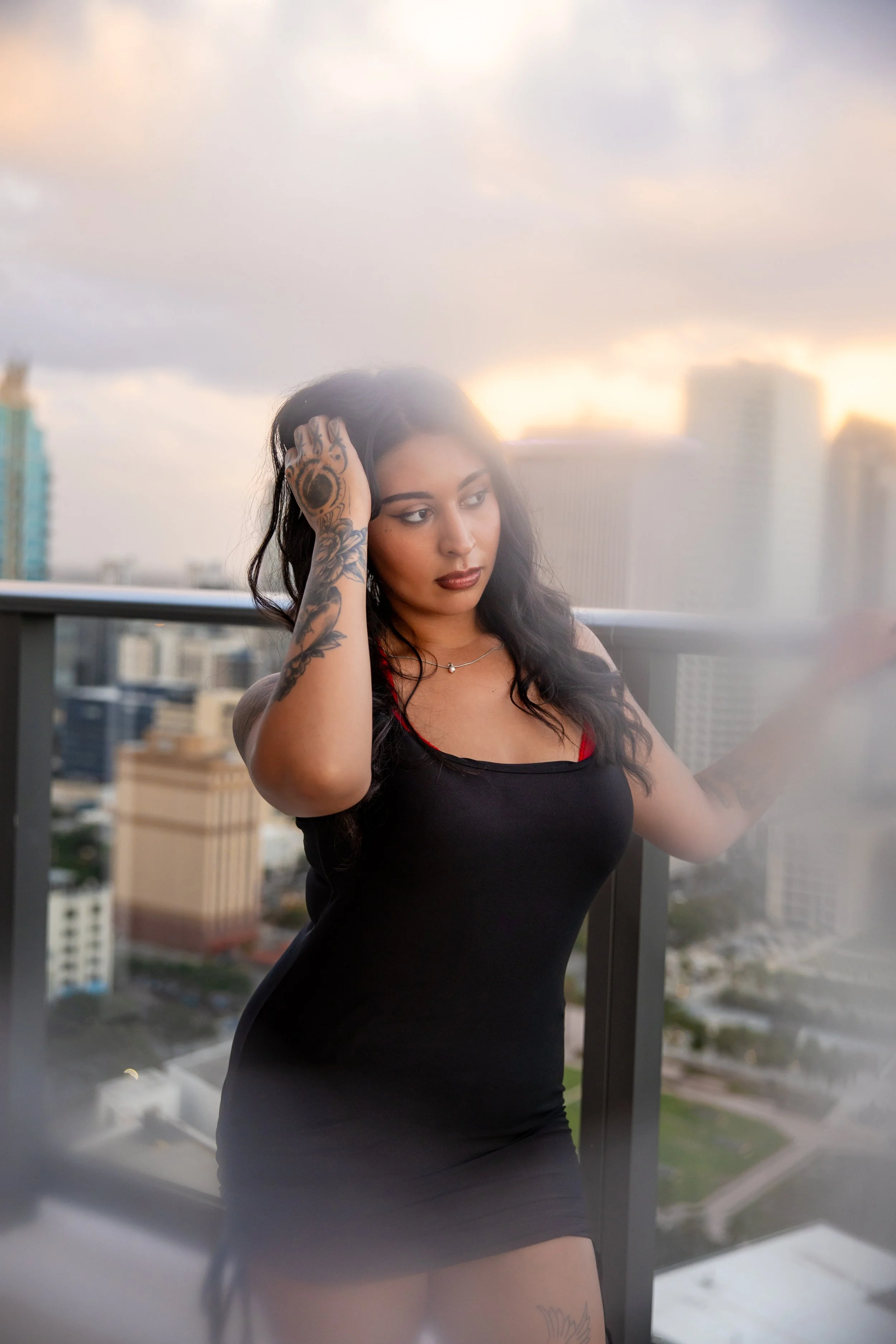 A woman with long black hair, tattoos on her arm, and wearing a black dress, standing on a balcony with a cityscape in the background during sunset.
