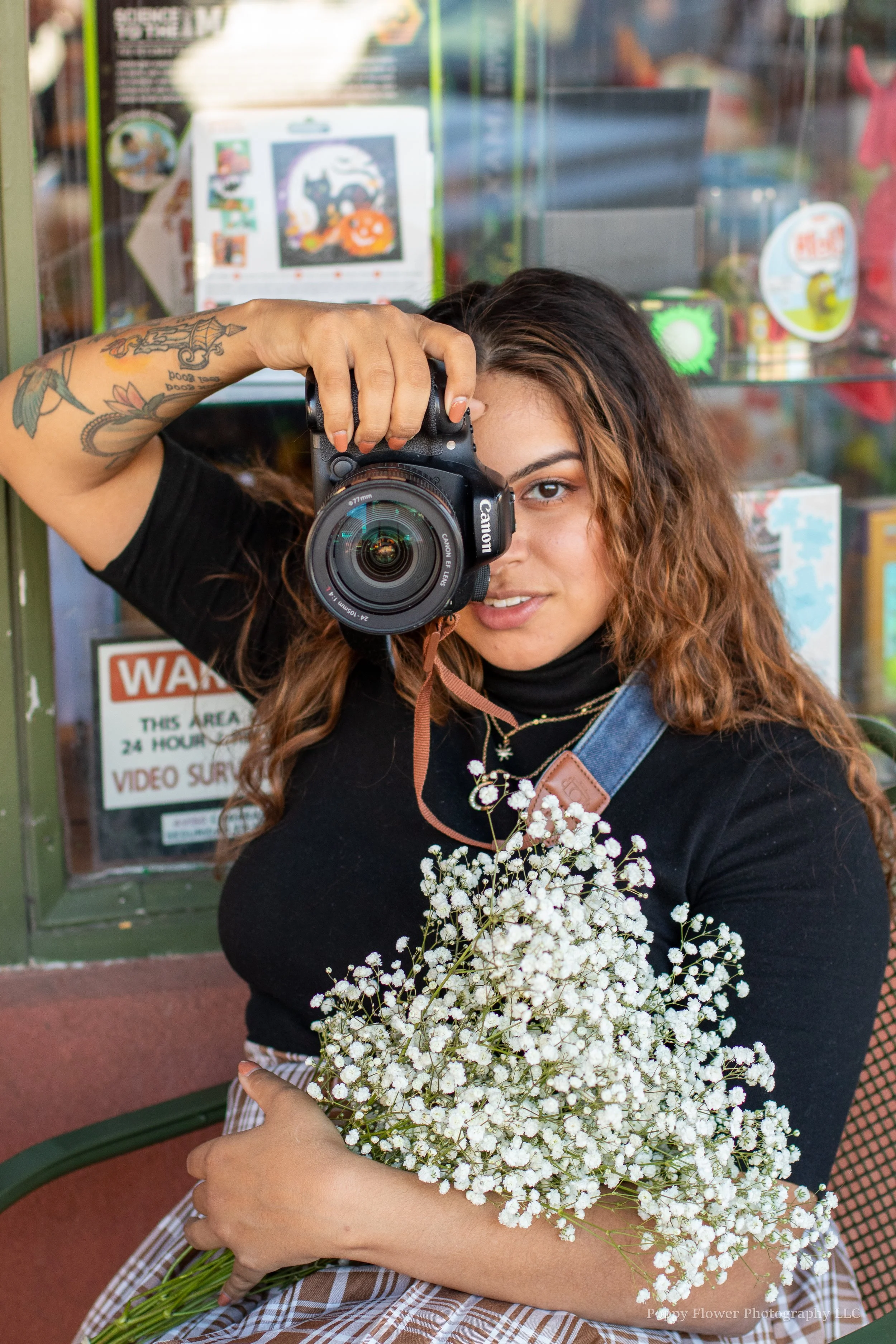 A woman with brown, curly hair and tattoos on her left arm holding a camera to her face and a bouquet of white baby’s breath flowers in front of her. She is wearing a black top and striped brown and white skirt, standing in front of a glass display case.