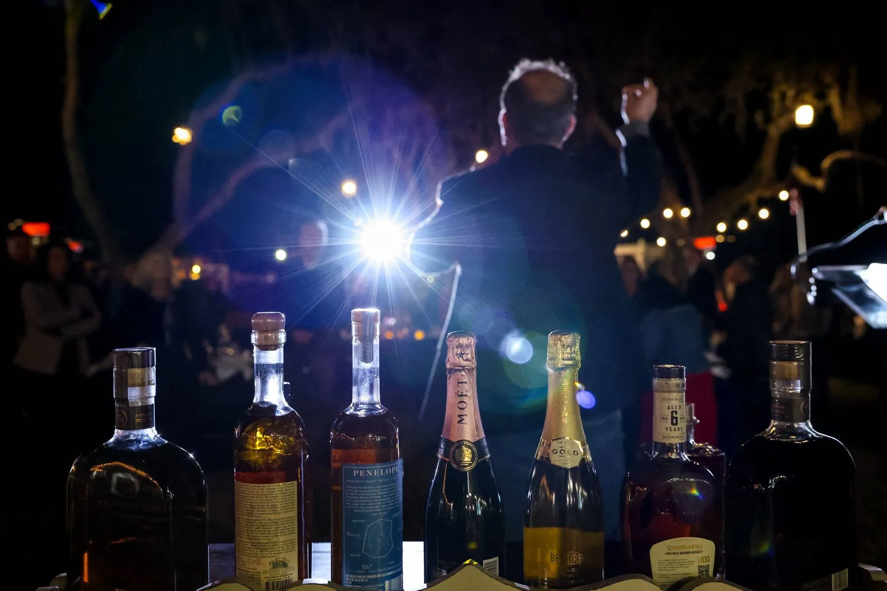 A person in a dark jacket giving a speech at an outdoor night event, with a spotlight in their back. Several bottles of liquor are in the foreground on a table, including whiskey, gin, and champagne, with a crowd of people in the background.