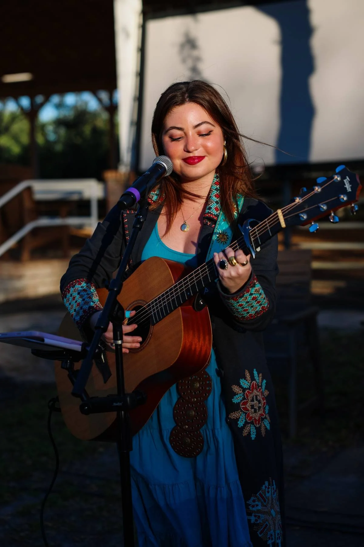 Young woman with brown hair, wearing a blue dress with embroidered patterns, playing an acoustic guitar and singing at an outdoor event during late afternoon or early evening.
