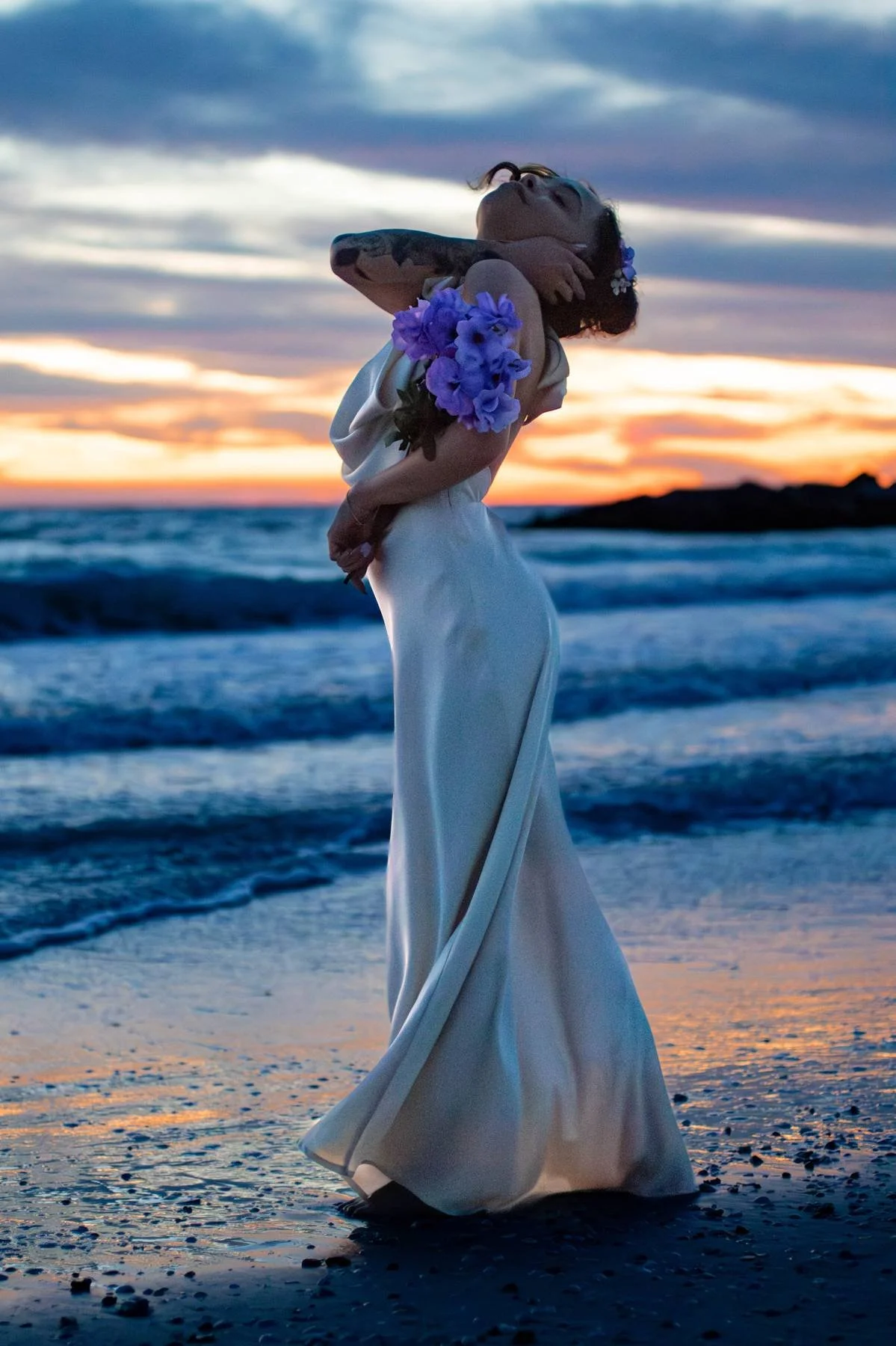 A woman in a long white dress with purple flowers on her arm, standing on a beach during sunset, with her head tilted back and arms around her neck.