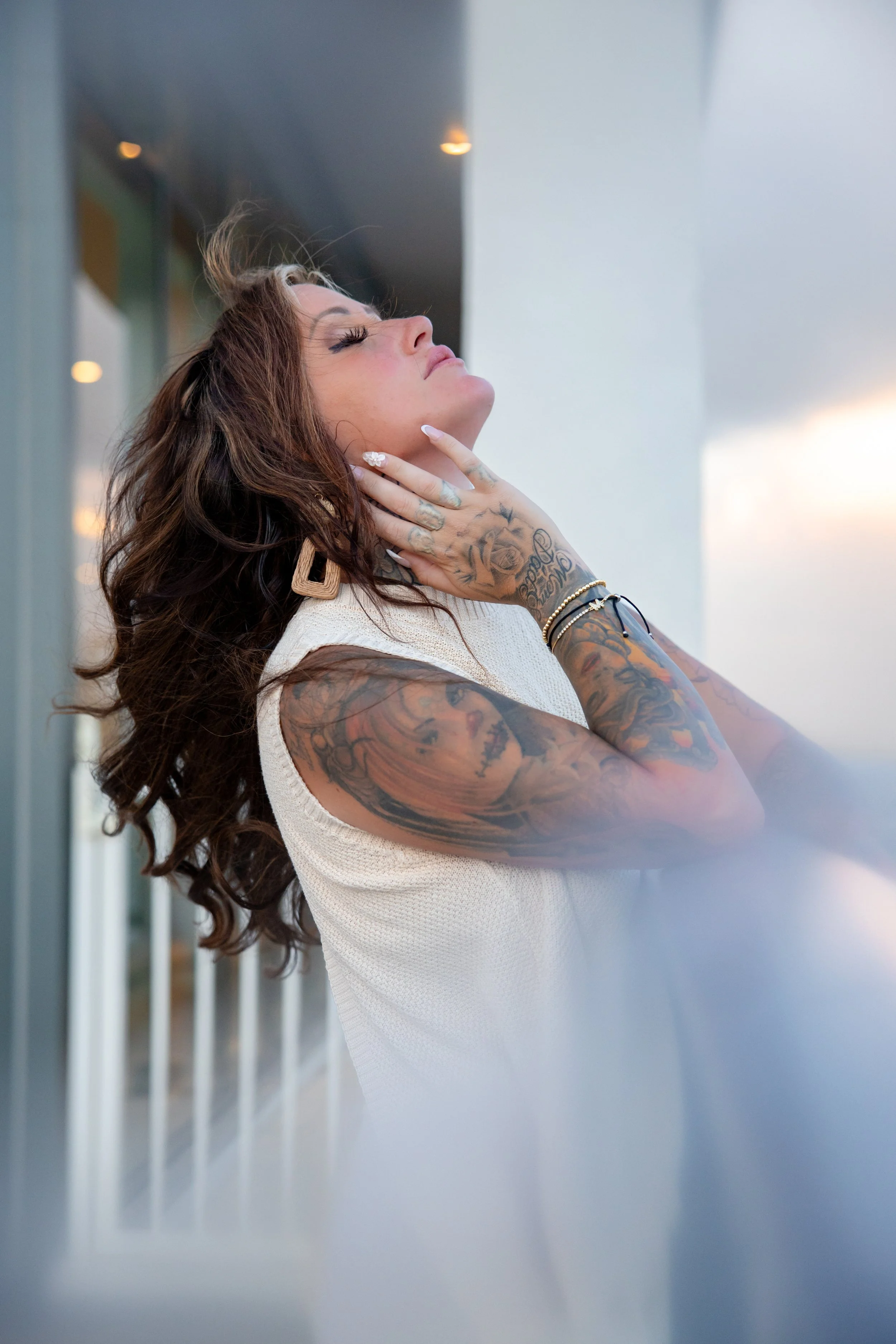 A woman with long curly brown hair, tattoos on her arms, and wearing earrings and bracelets poses with her eyes closed and hand on her neck, outdoors near a glass building.