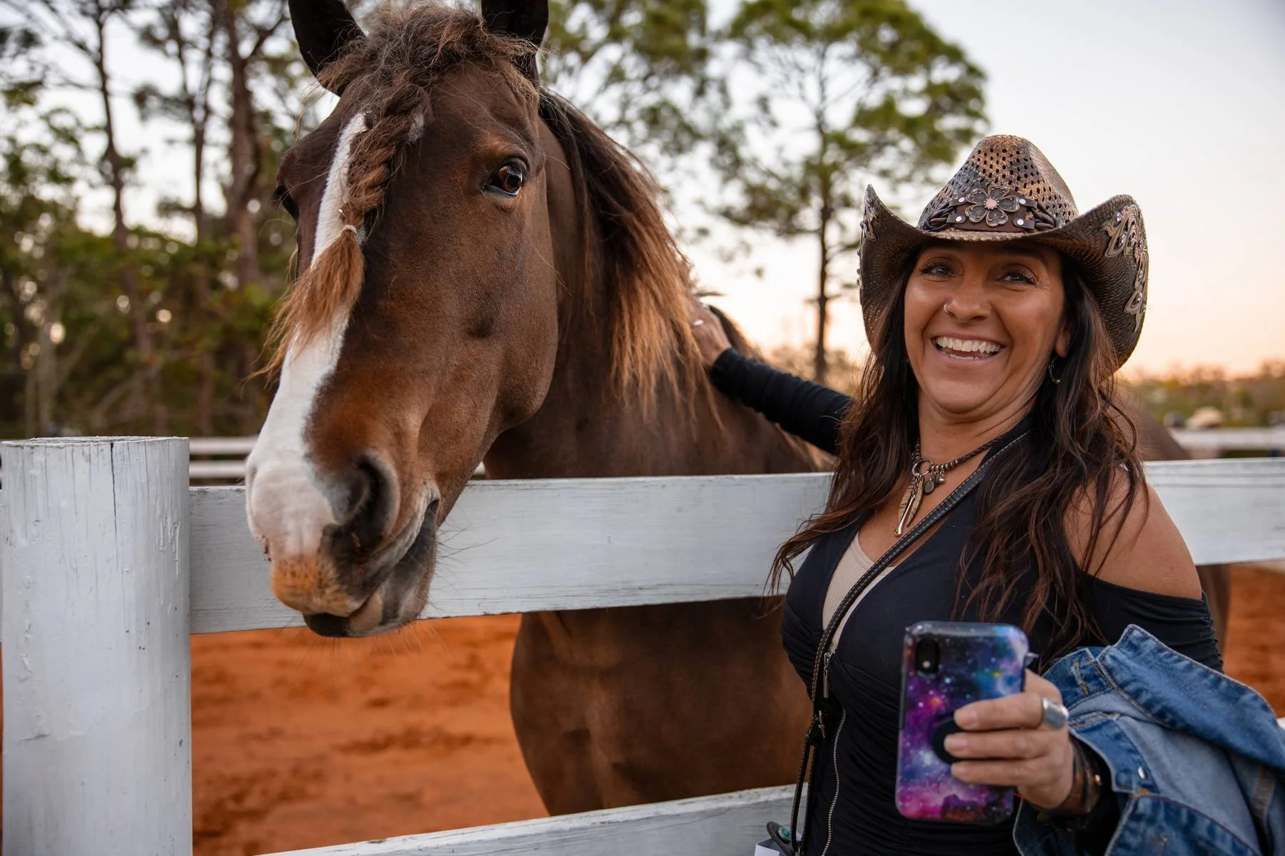 A smiling woman with dark hair wearing a cowboy hat and earrings taking a selfie with a horse at a farm or ranch during sunset.