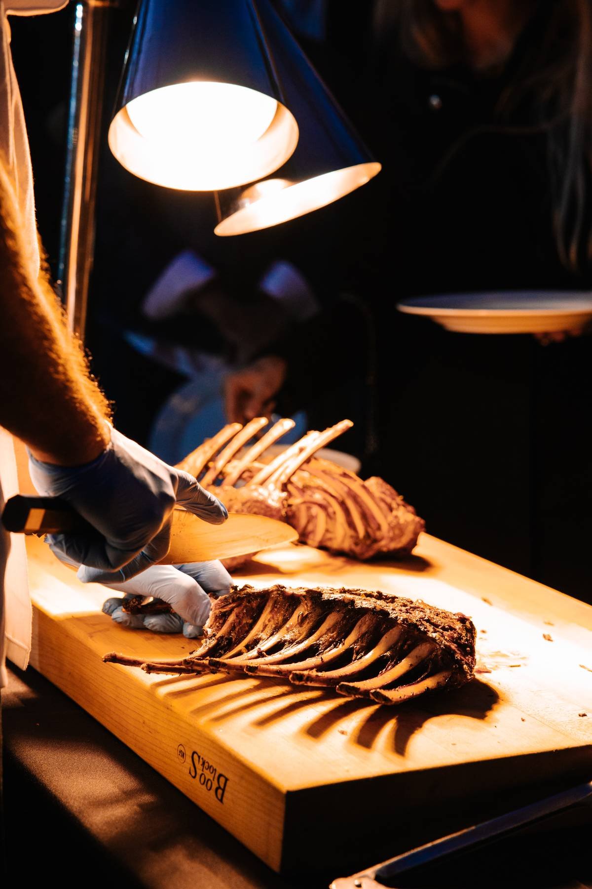A chef carving a large rack of cooked ribs on a wooden cutting board under a bright lamp.