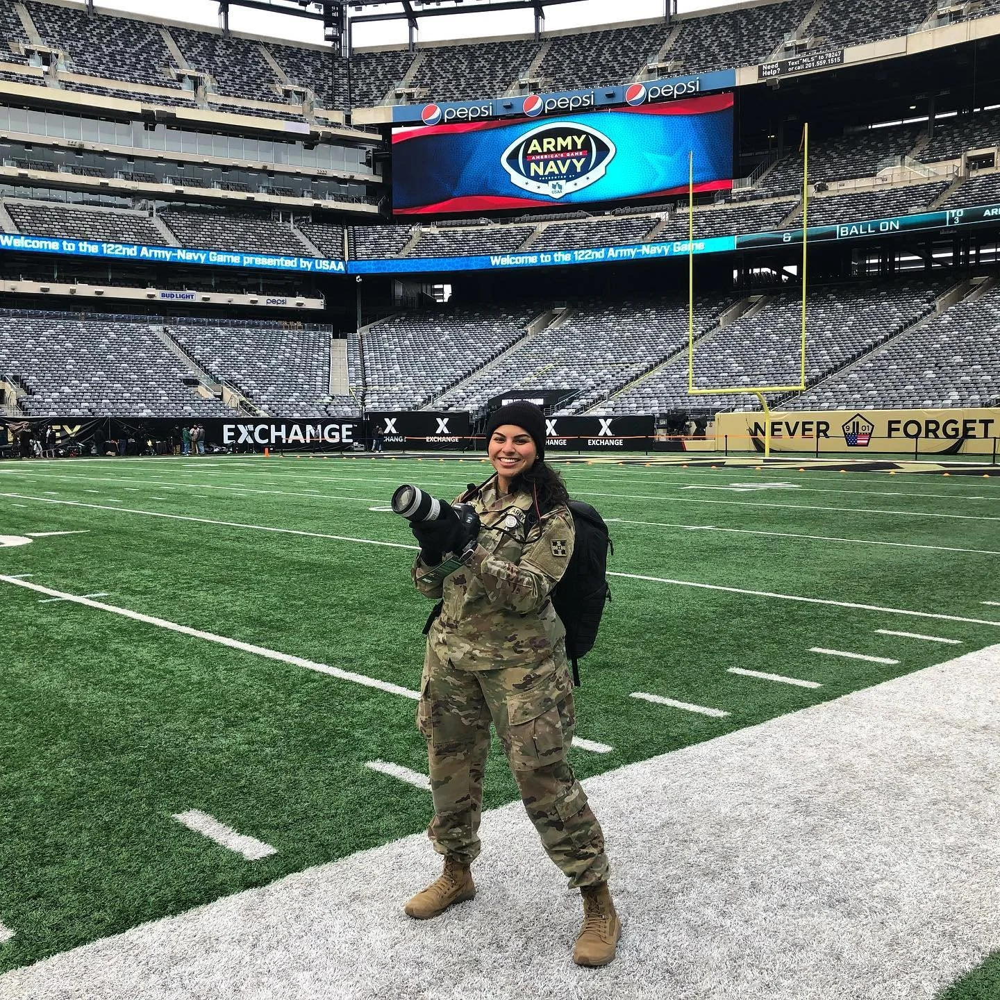 A woman in military camouflage uniform standing on a football field, smiling and holding a camera with a large lens. The stadium seats are mostly empty, and there are banners and electronic signs in the background.