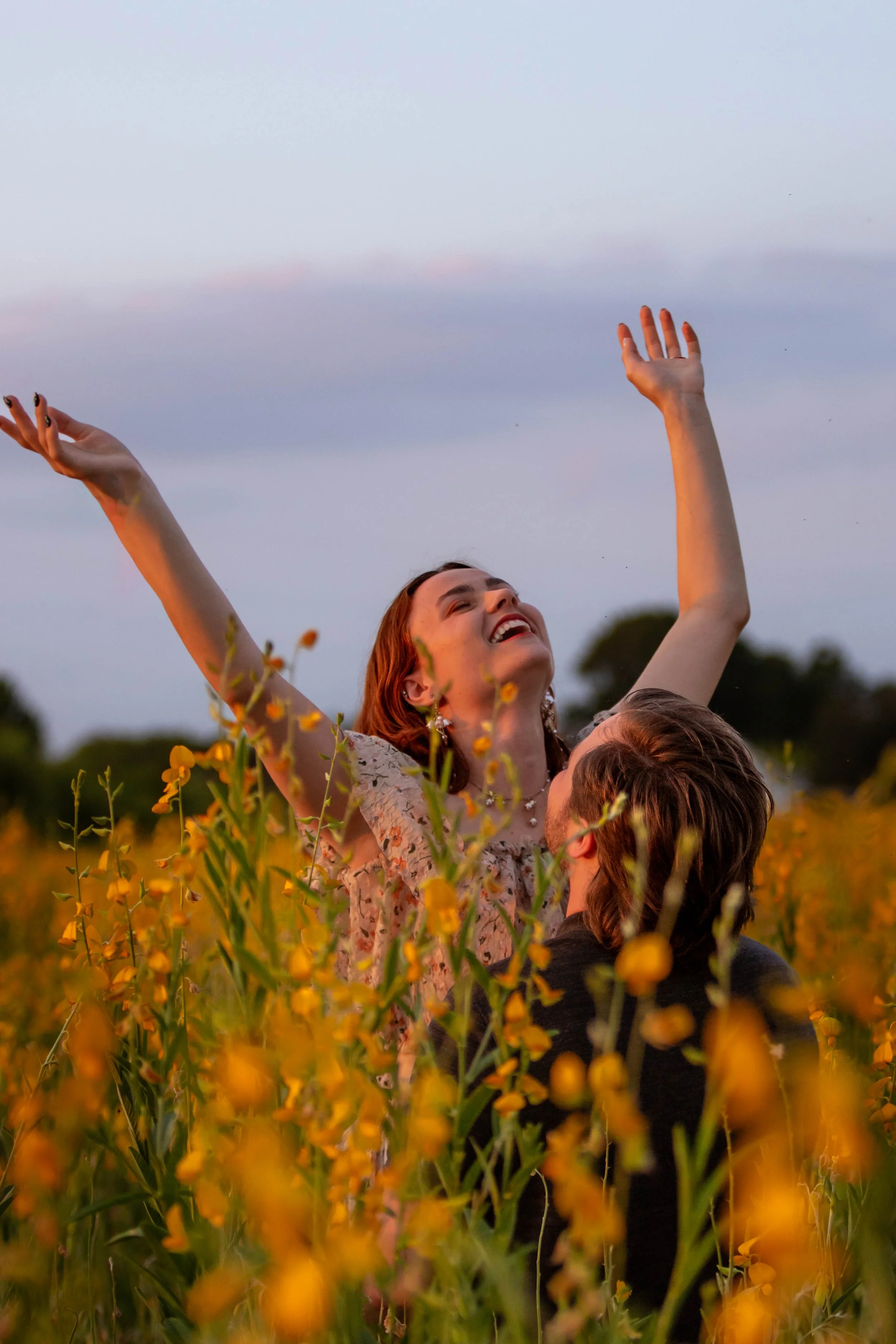 Young woman with red hair smiling with eyes closed, raising arms in a yellow flower-filled field during sunset or dusk.