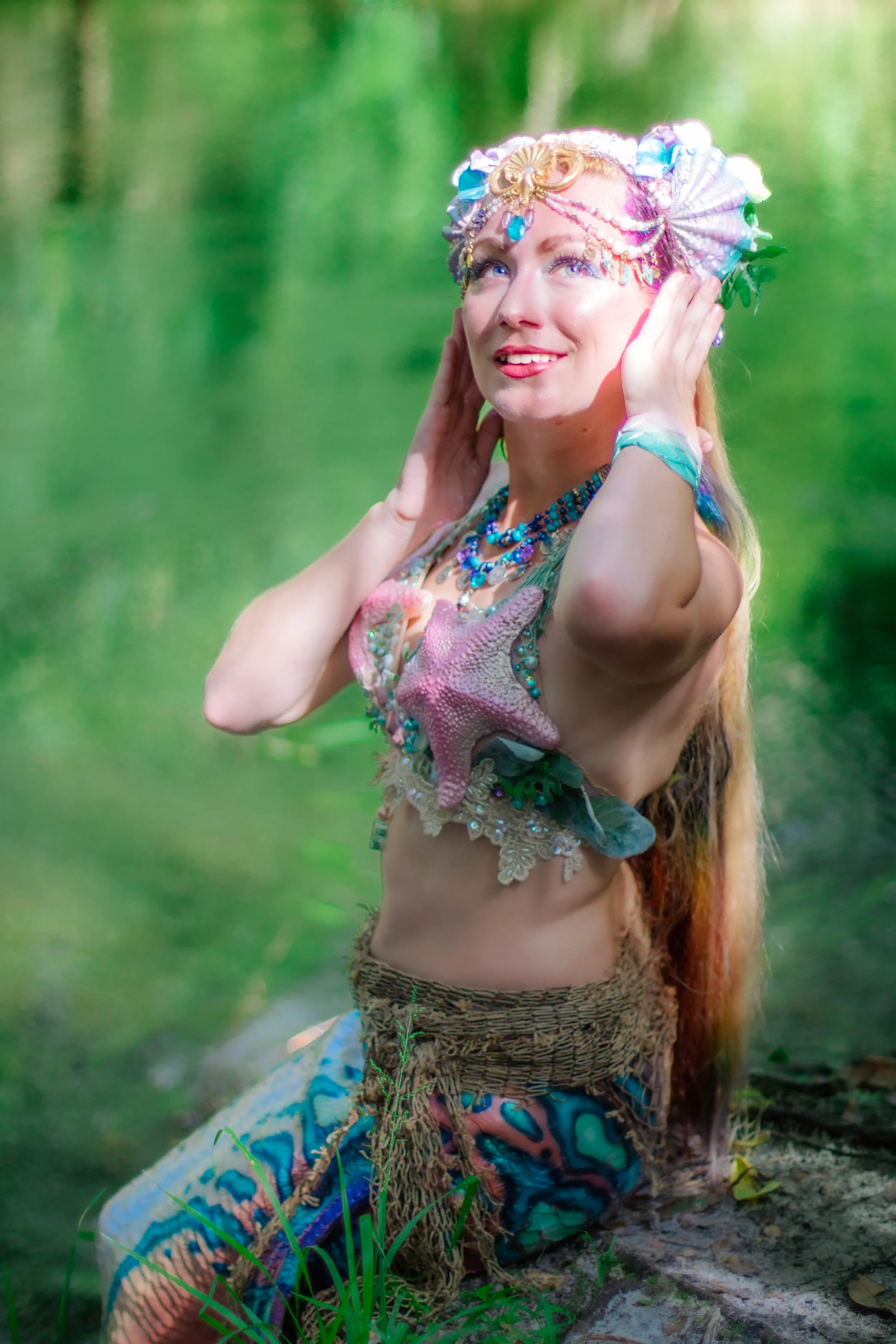 A woman with long, reddish hair and blue eyes dressed in a mermaid costume with sea shell and starfish accessories, posing outdoors near a body of water with green trees in the background.