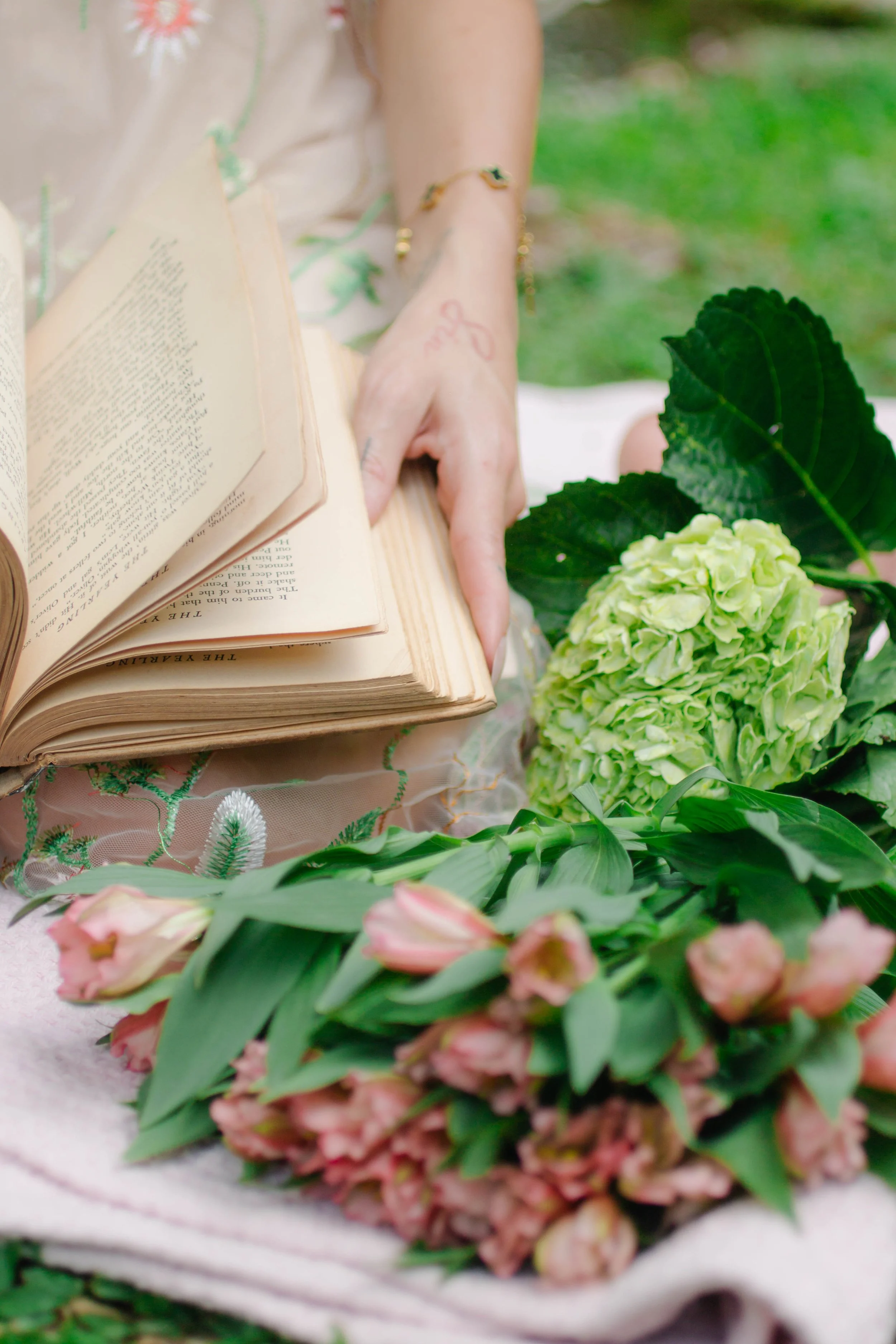 Person holding an open book and surrounded by various flowers, including green hydrangeas and pink blossoms, with a blurred green background.