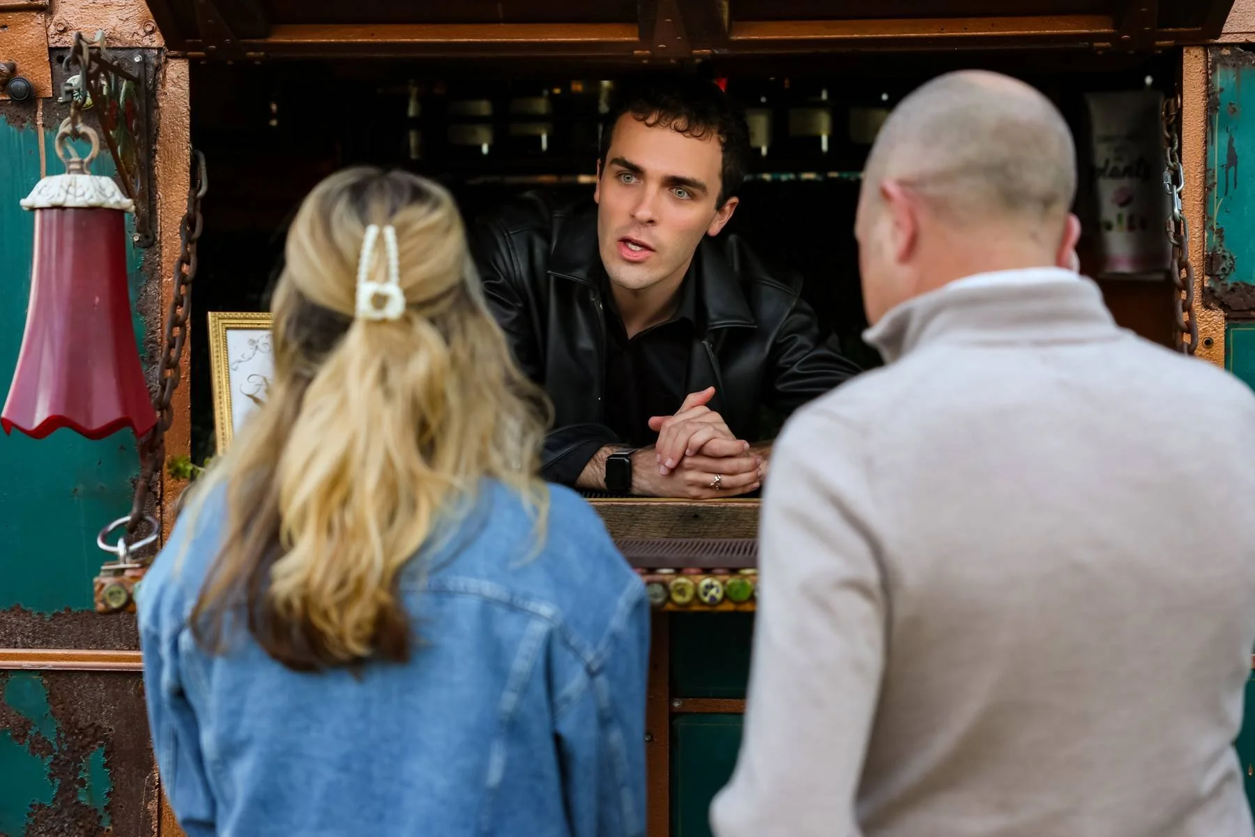 A man with short dark hair and a black leather jacket talking to a blonde woman with a hair clip and a man with a shaved head and glasses at a rustic booth, possibly in a market or fair.