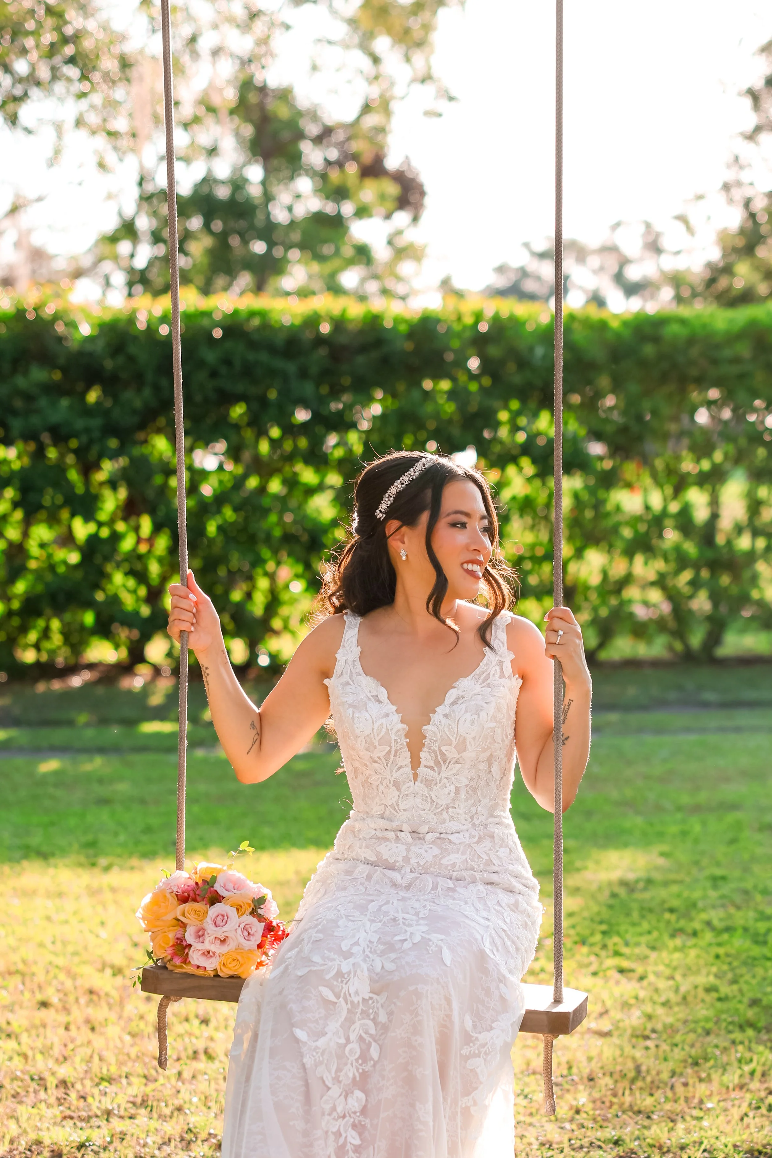 A bride wearing a white lace wedding dress, sitting on a swing outdoors, holding a bouquet of pink, peach, and cream roses. She has dark hair styled with a pearl headband and is smiling, with greenery and trees in the background.