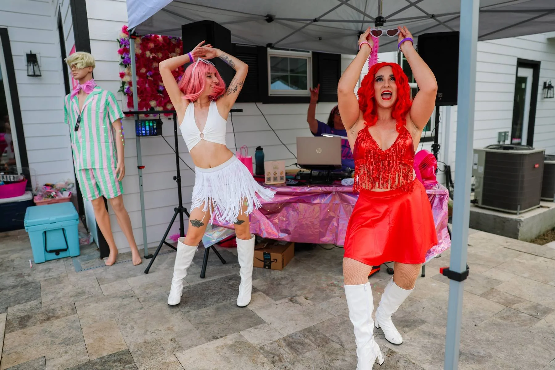 Three women dancing at an outdoor party under a canopy, with a decorated backdrop and a DJ setup in the background. One woman has bright red hair and is wearing a red outfit, another has pink hair and a white fringe outfit, and the third has blonde h