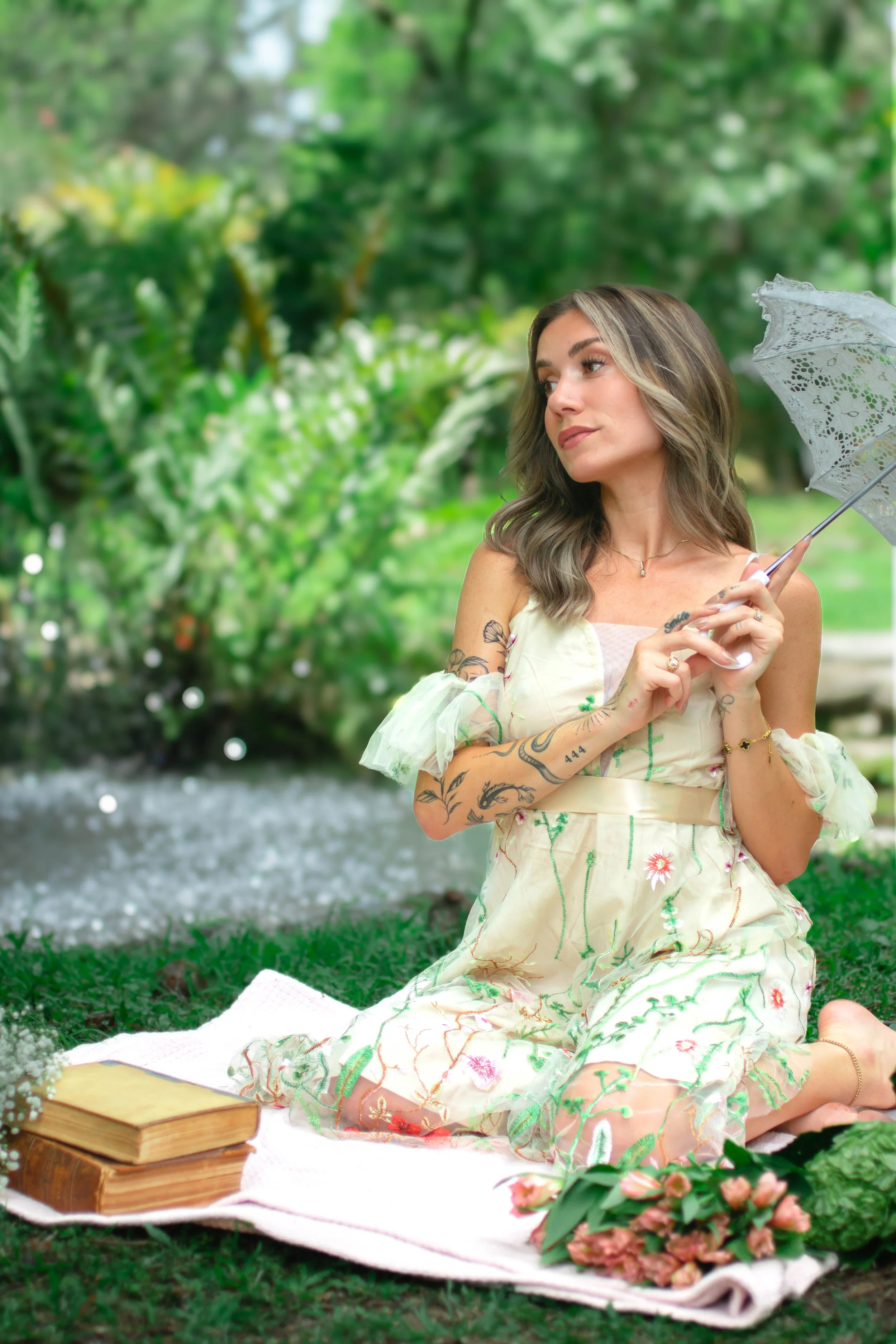 A woman sitting on a blanket outdoors holding a lace parasol, surrounded by greenery and flowers, with books and a floral arrangement nearby.