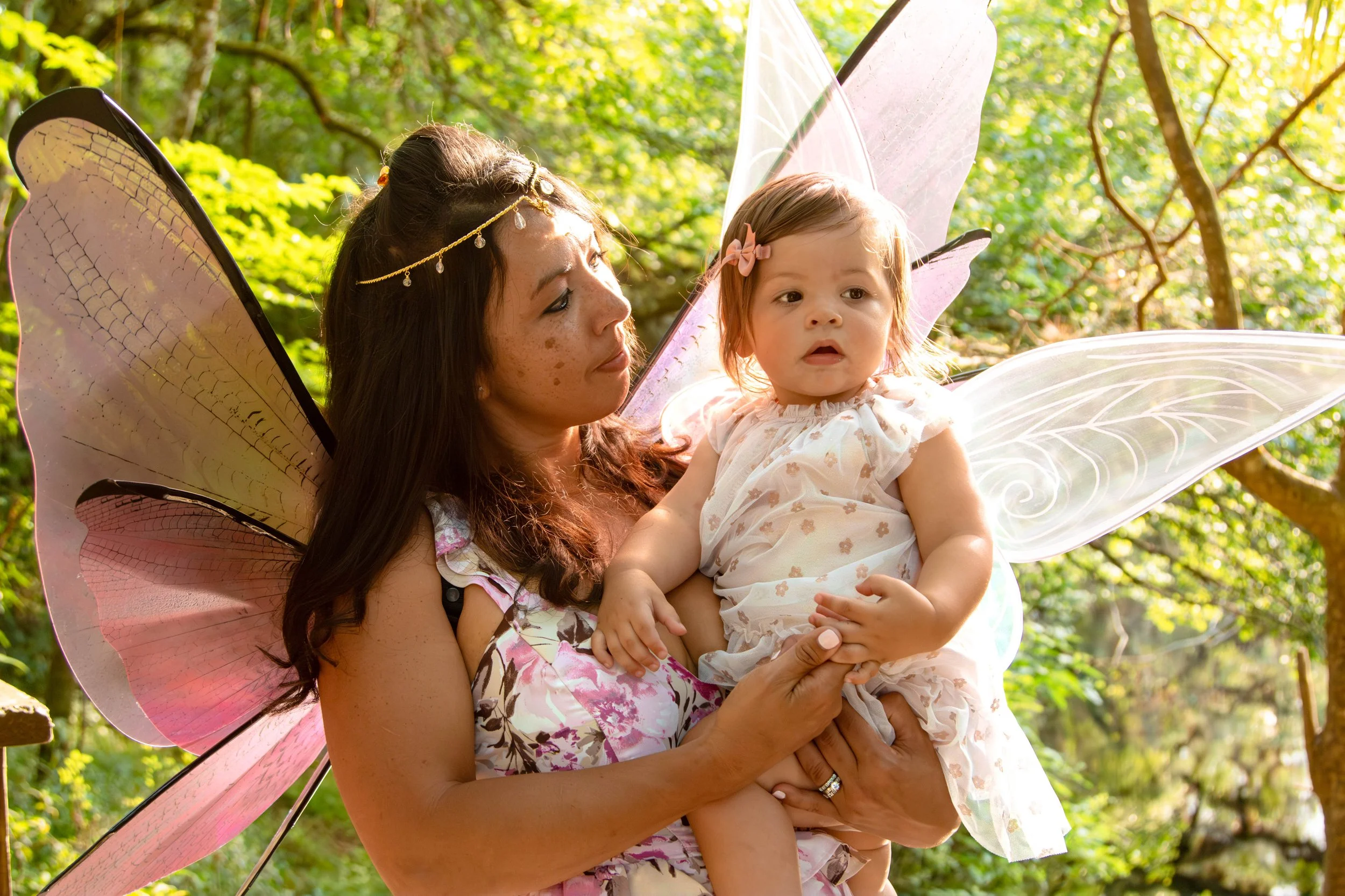 A woman with fairy wings holding a young girl with fairy wings in a wooded outdoor setting.