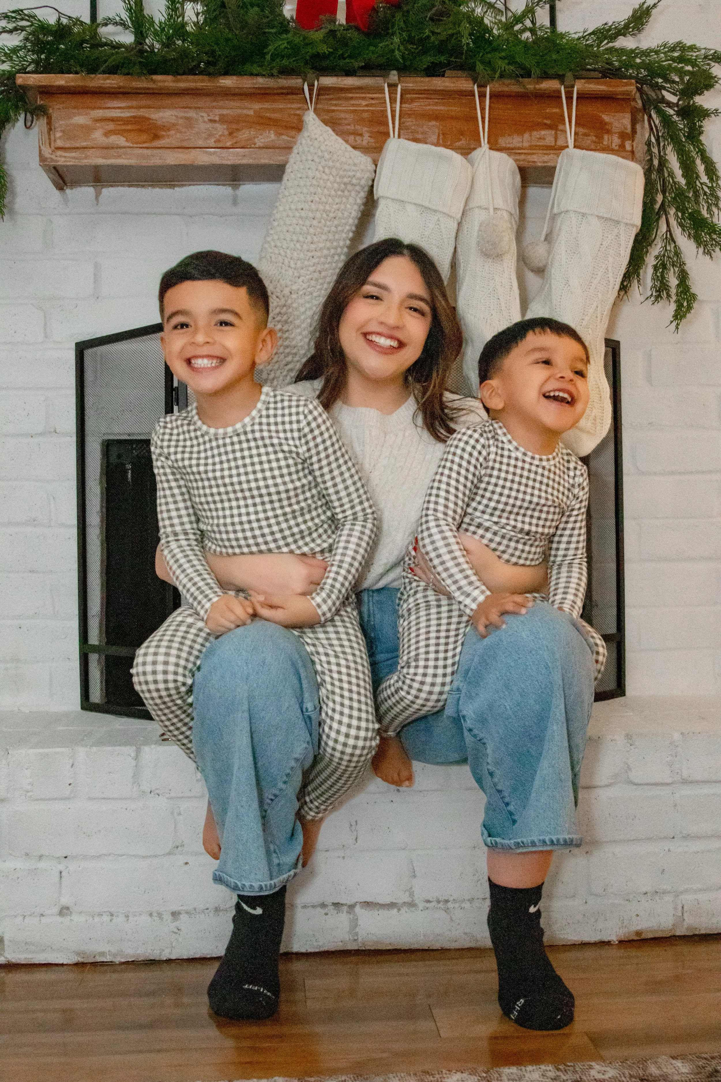 A woman and two young boys sitting on a chair in front of a fireplace with white stockings hanging above, smiling.