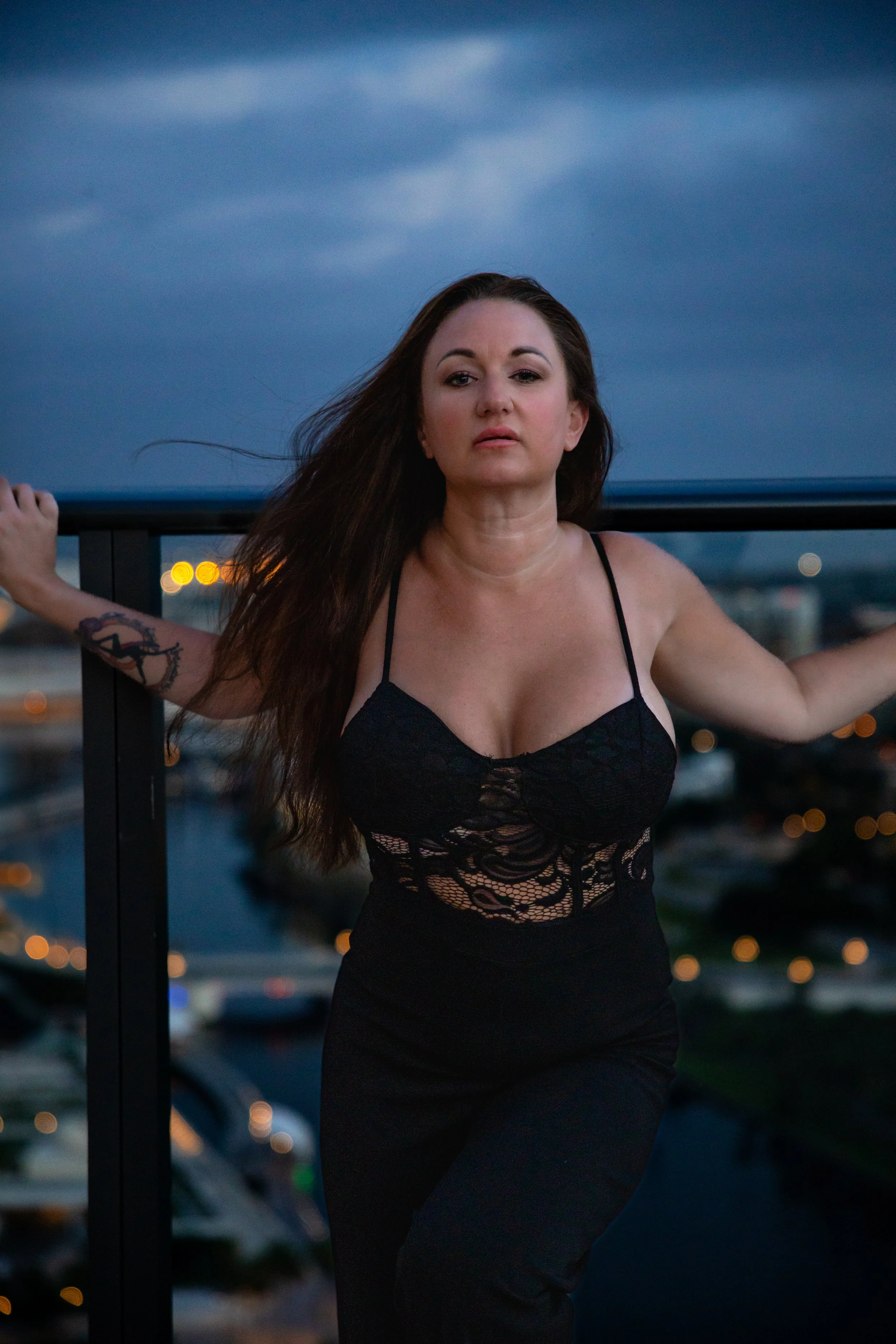 A woman with long brown hair and tattoos on her arm, wearing a black lace dress, standing on a rooftop with a cityscape and cloudy sky in the background at dusk.