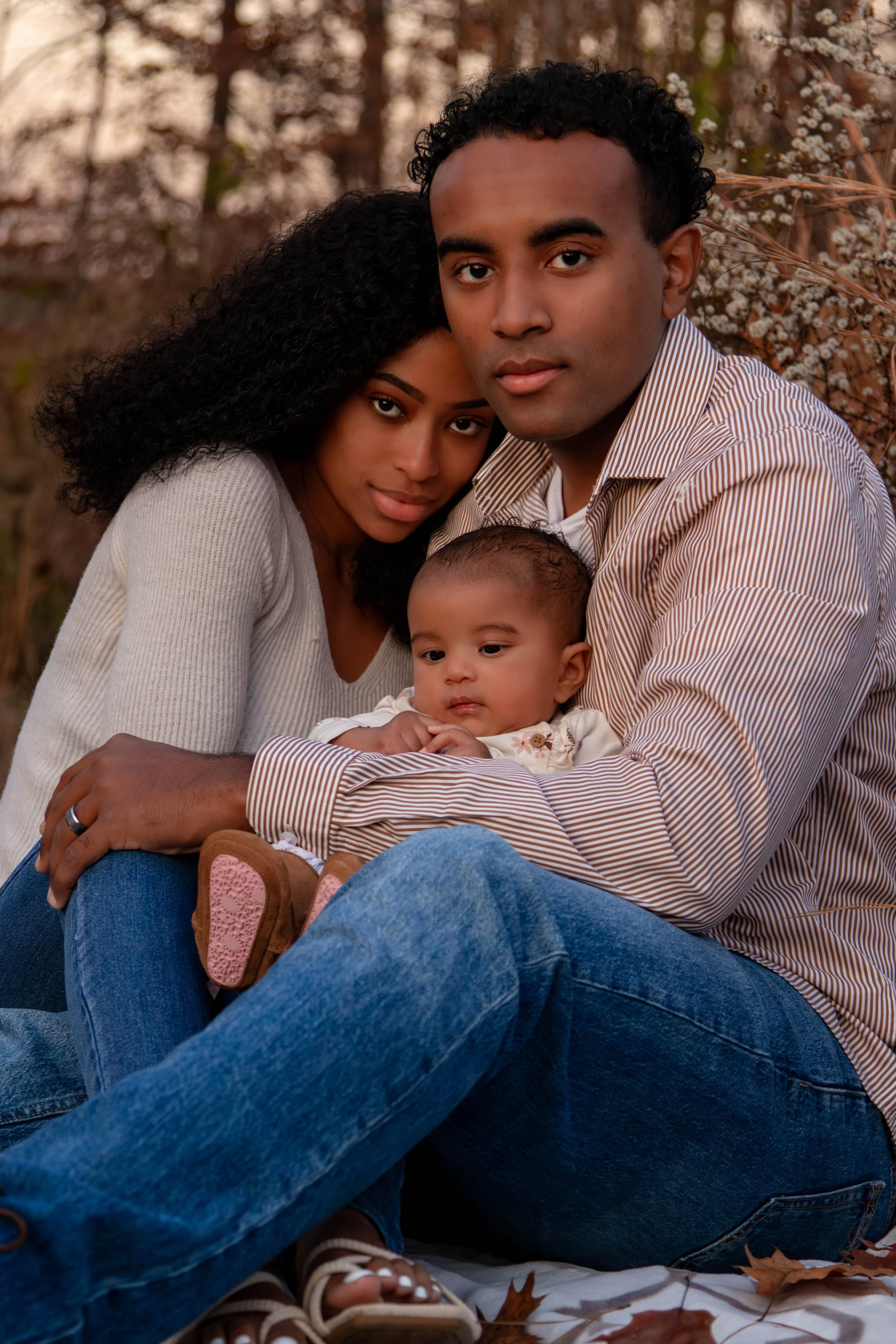 A family of three sitting outdoors in a wooded area with white blossoms and fallen leaves, posing for a photo. The woman has curly black hair and is wearing a light sweater, the man has short curly hair and is wearing a striped shirt, and the child i