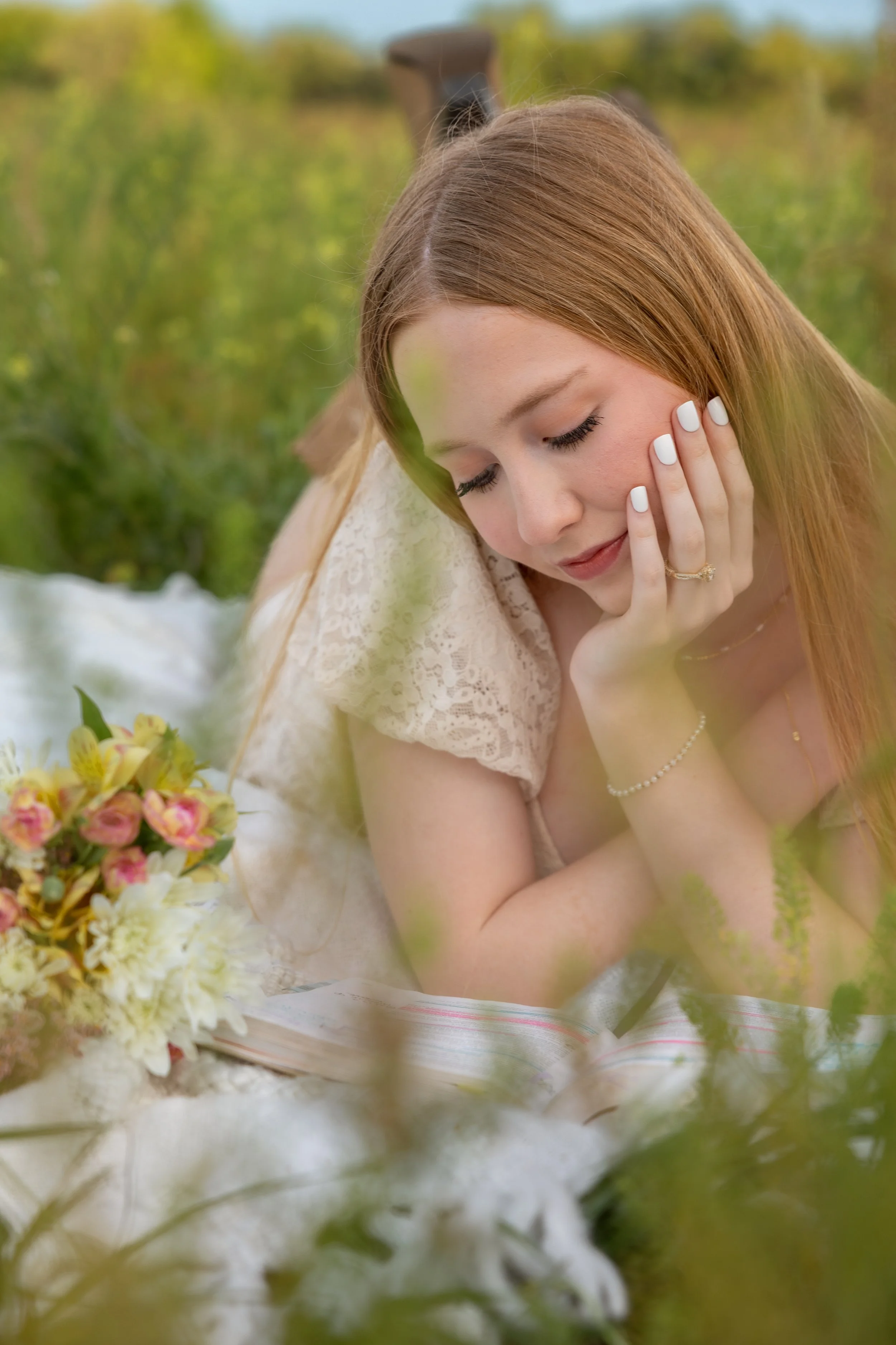 A young woman with long red hair, closed eyes, and a gentle expression, resting her face on her hand outdoors with greenery and flowers around her.