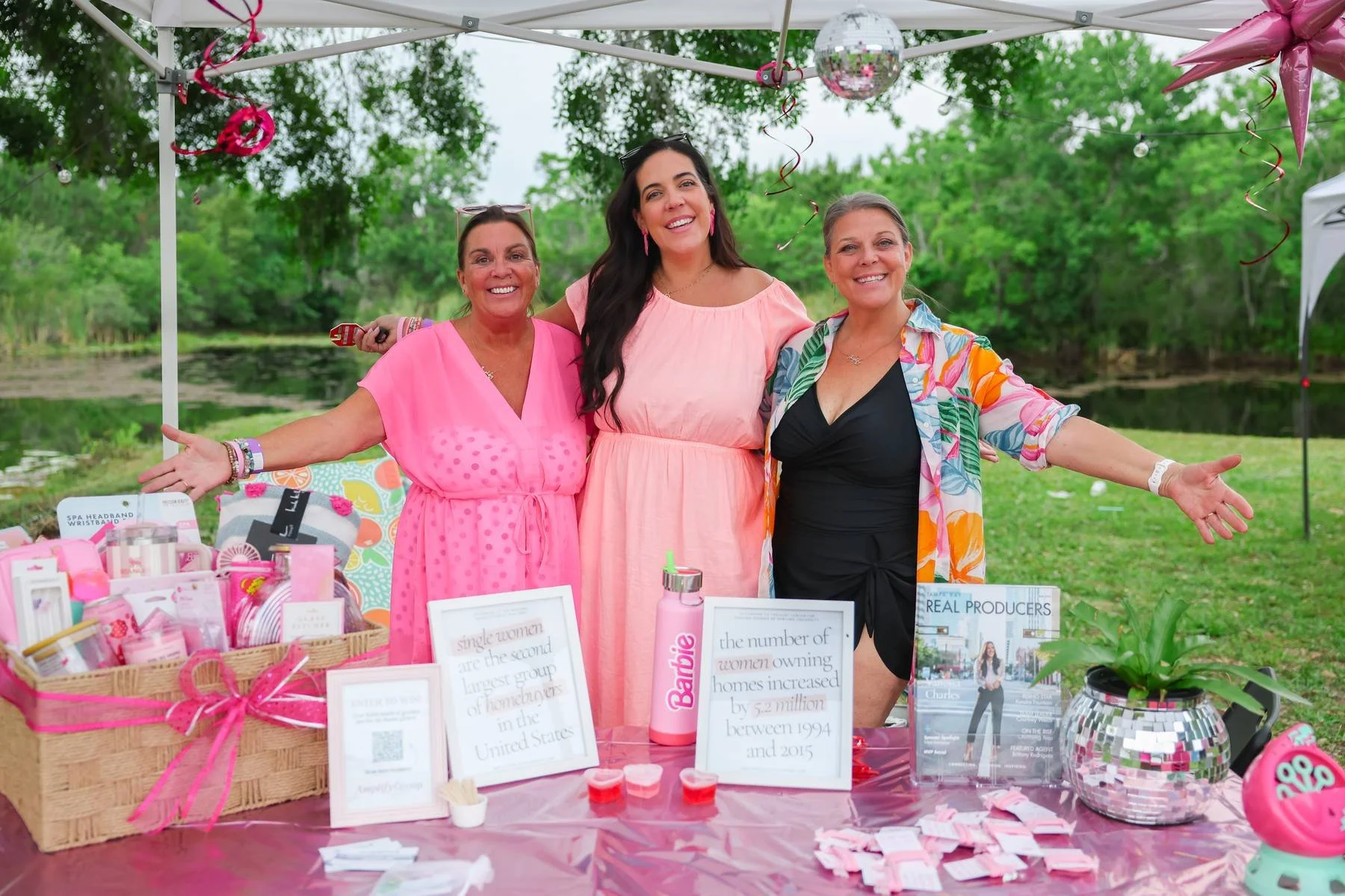 Three women standing behind a pink table decorated with pink ribbons and items, at an outdoor event with green trees in the background. They are smiling and wearing colorful dresses, with various informational signs and products on the table.