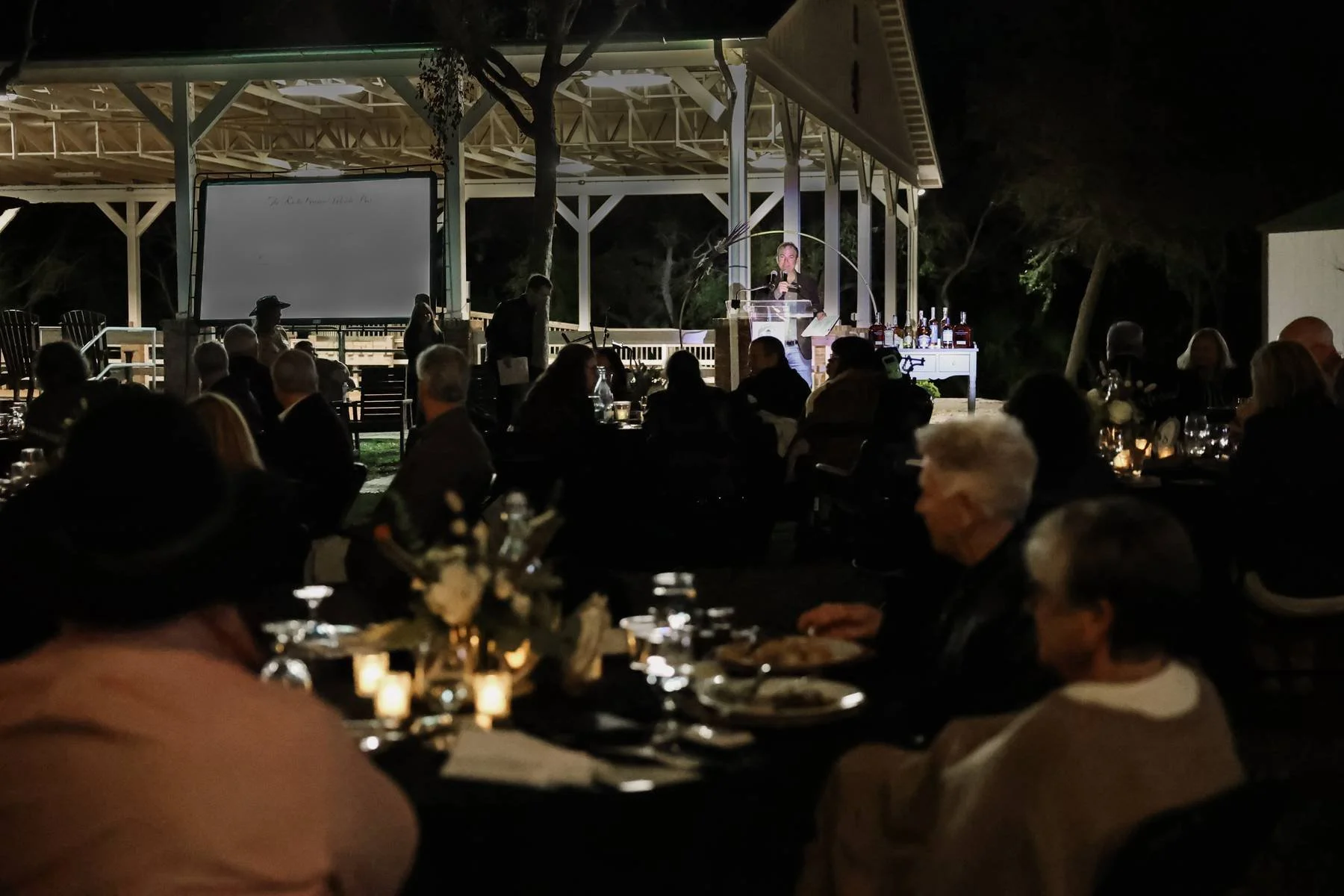 Nighttime outdoor event with people seated at tables, listening to a speaker on a stage, with a whiteboard and bottles of alcohol nearby, under a covered pavilion surrounded by trees.