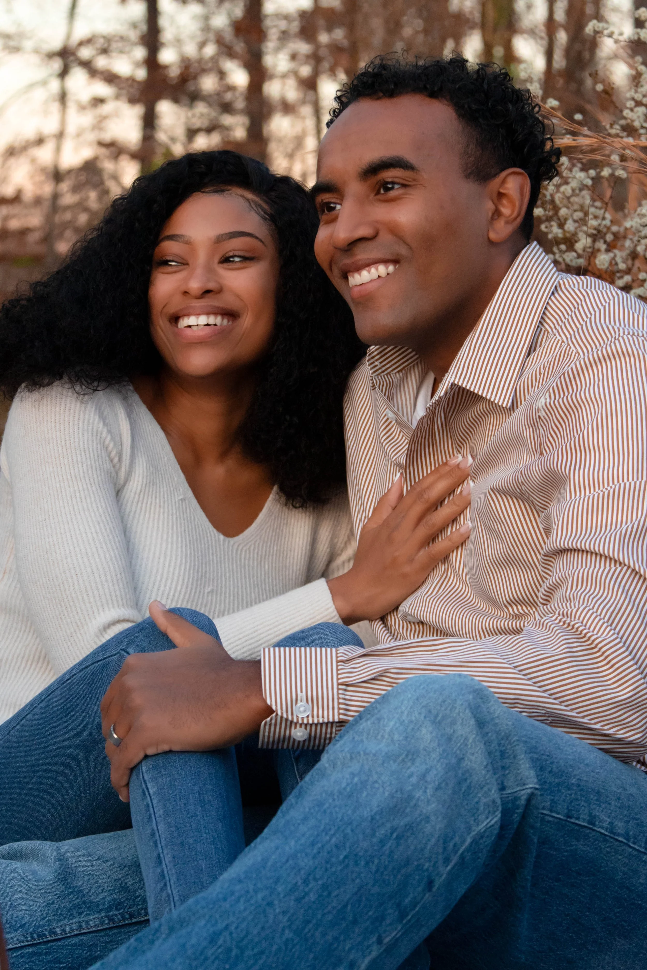 A smiling couple sitting outdoors in a park surrounded by blooming trees, enjoying each other's company.