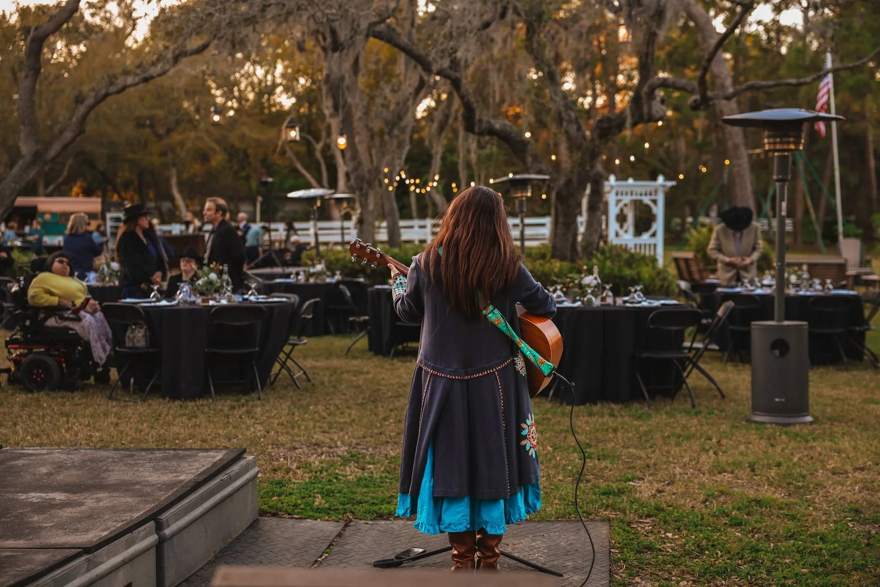 A person with long brown hair playing an acoustic guitar outdoors at an evening event, surrounded by tables with black tablecloths and guests, trees with string lights, and heaters in the background.