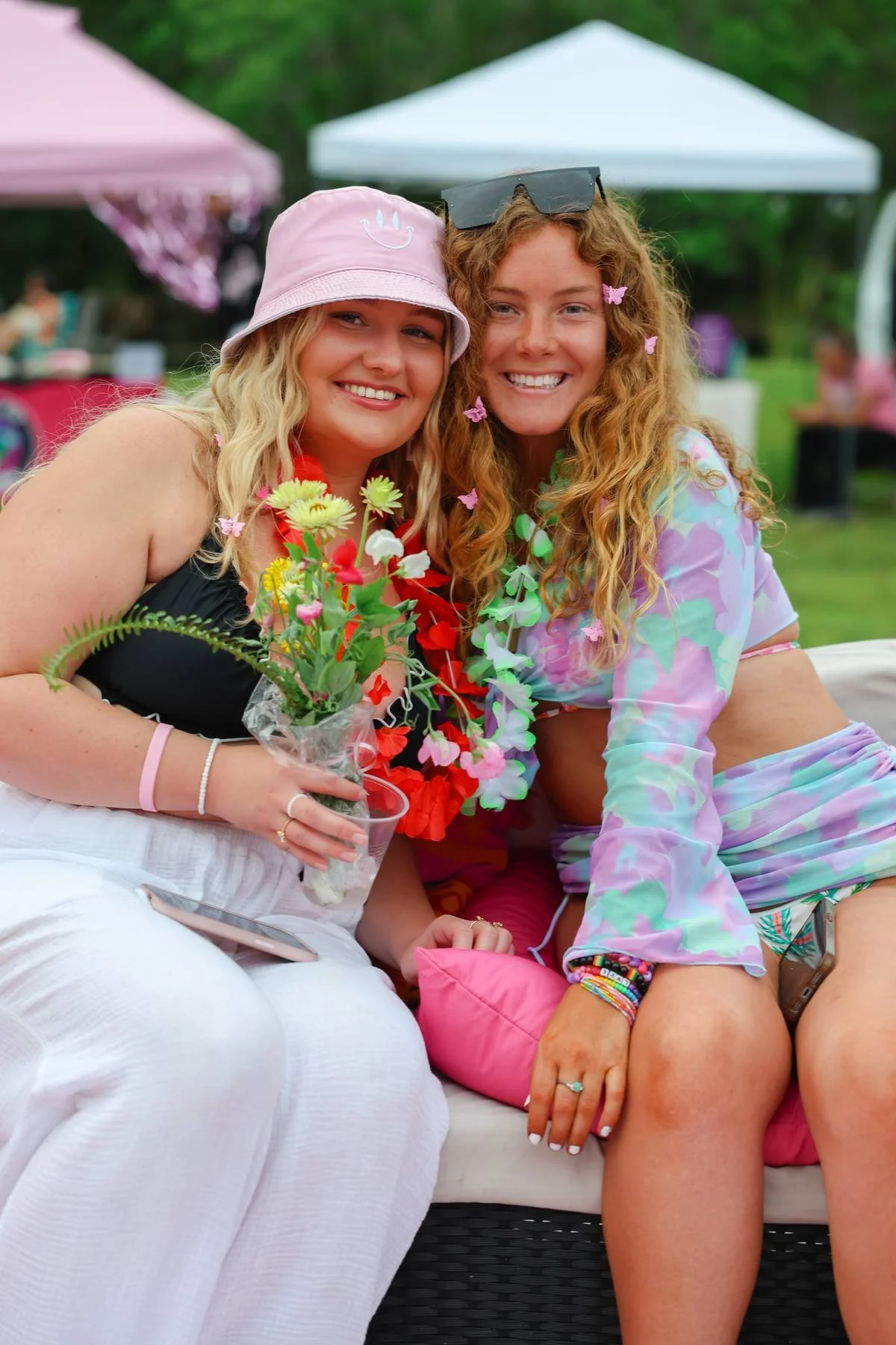 Two young women sitting close together outdoors at a celebration or gathering, smiling at the camera. One woman is holding a small bouquet of flowers. They are dressed casually with colorful accessories, and there are tents and other people in the ba