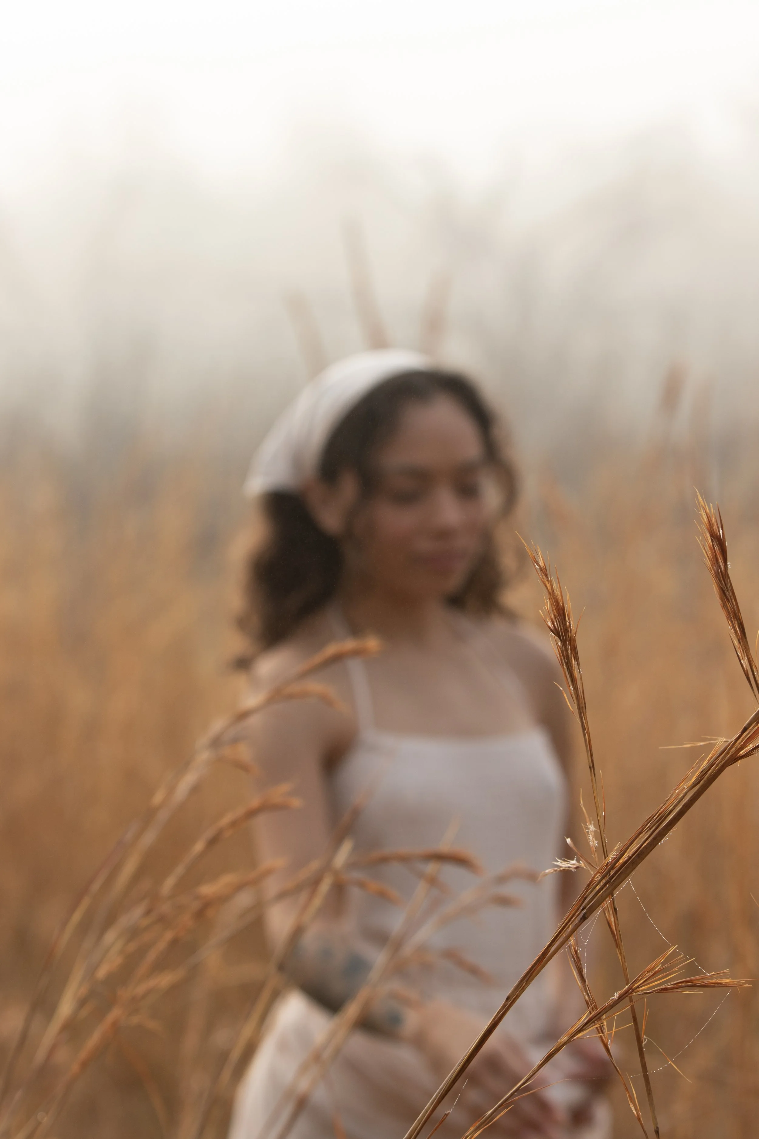 A woman with dark hair, glasses, and a white top, standing in a field of tall, dried grasses. The background is blurred, emphasizing the grasses in the foreground.
