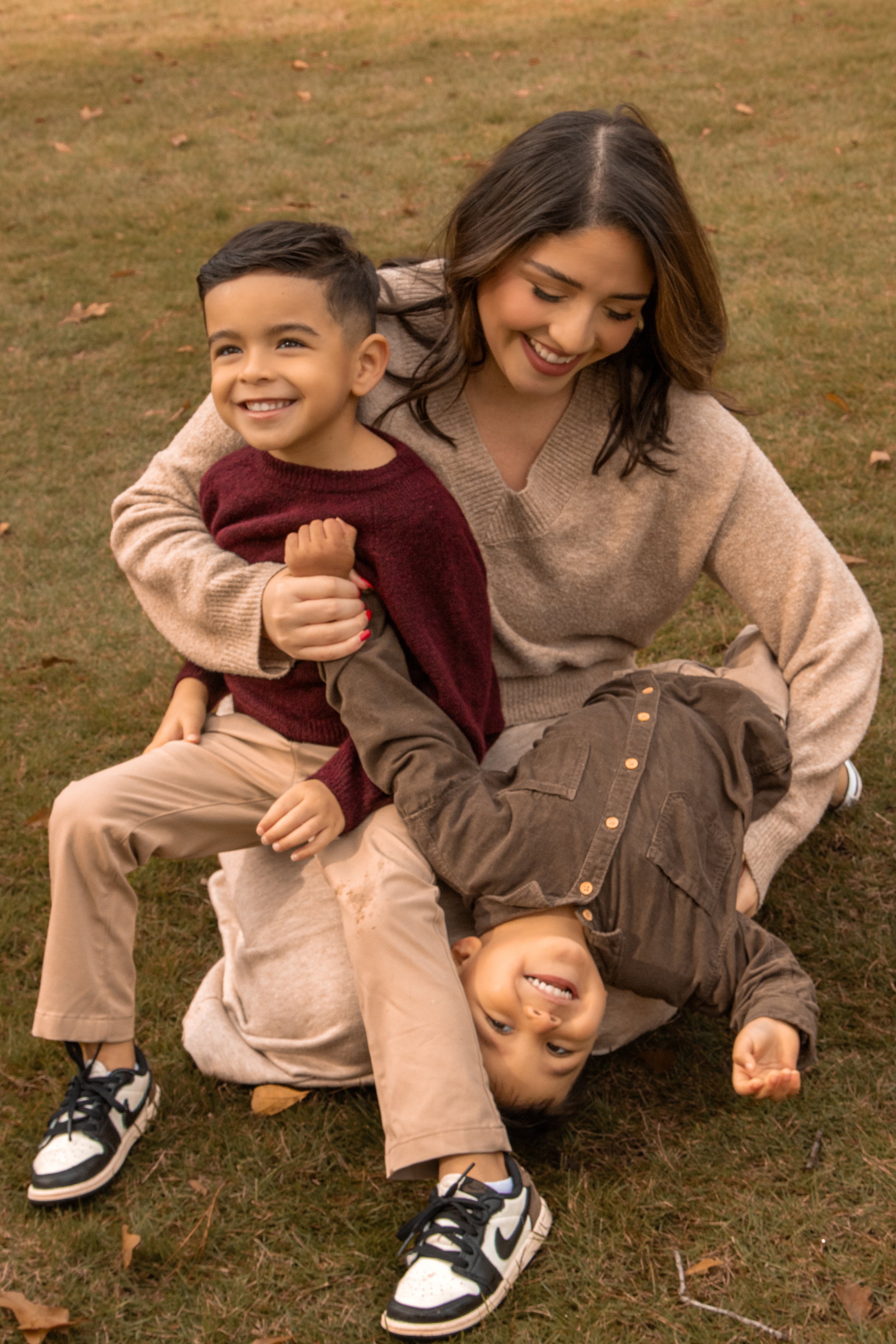 A woman and two young boys playing on the grass outside. One boy is reclining upside down, and the other boy is sitting on the woman, who is smiling and laughing.