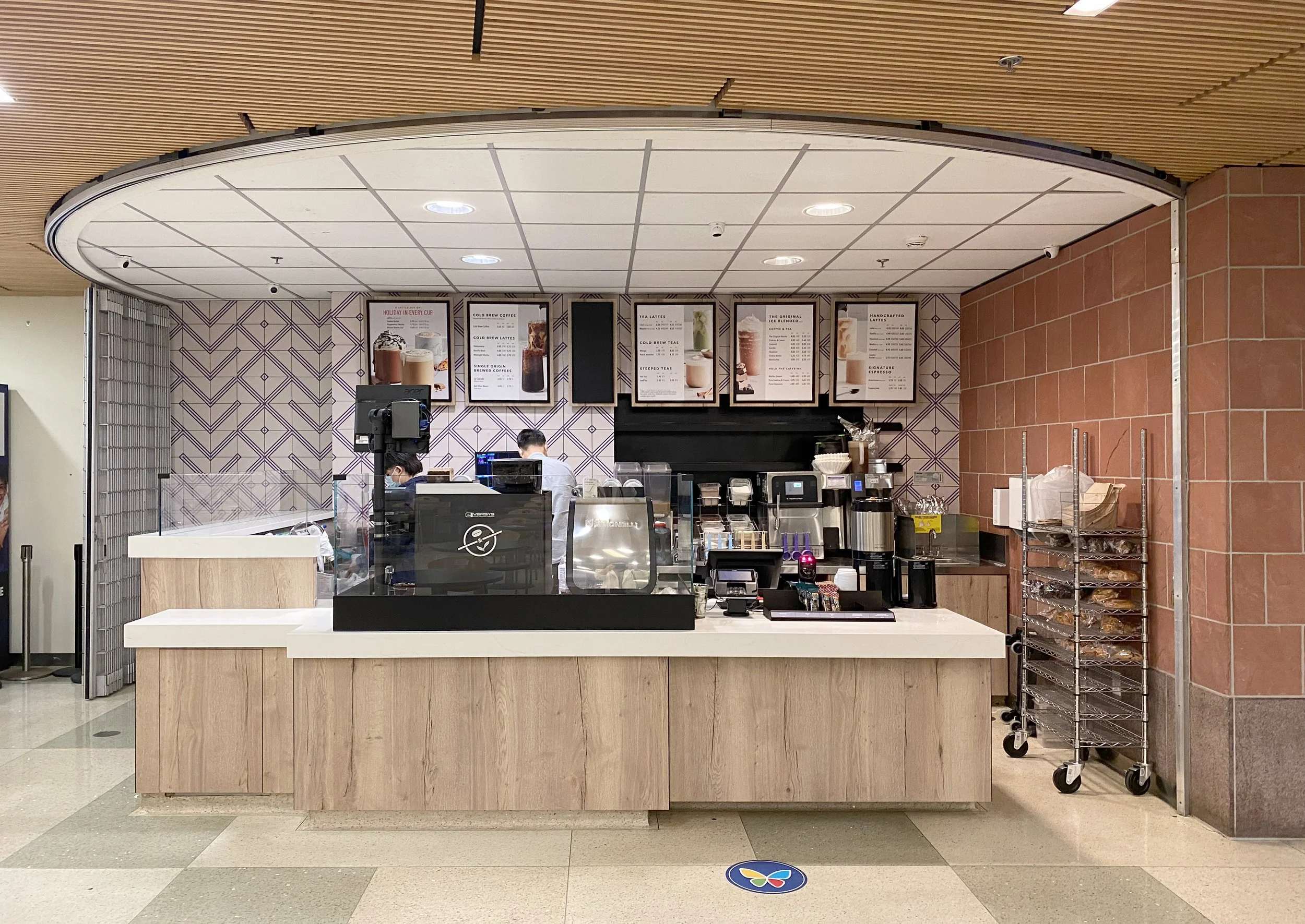 A coffee shop counter with menu signs overhead, staff behind the counter, and a metal rolling cart on the right with a few items on it. The ceiling has a combination of wood and white tiles, and the wall behind the counter has a geometric pattern.