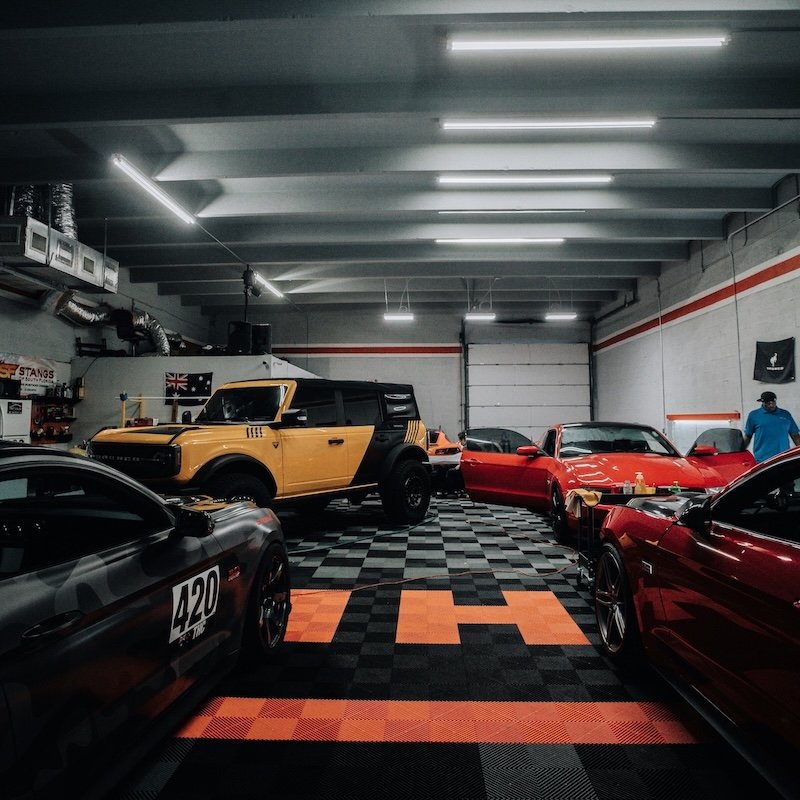 Multiple luxury cars and a yellow off-road vehicle inside a garage with checkered and orange flooring, white walls, fluorescent lighting, and a person in a blue shirt.