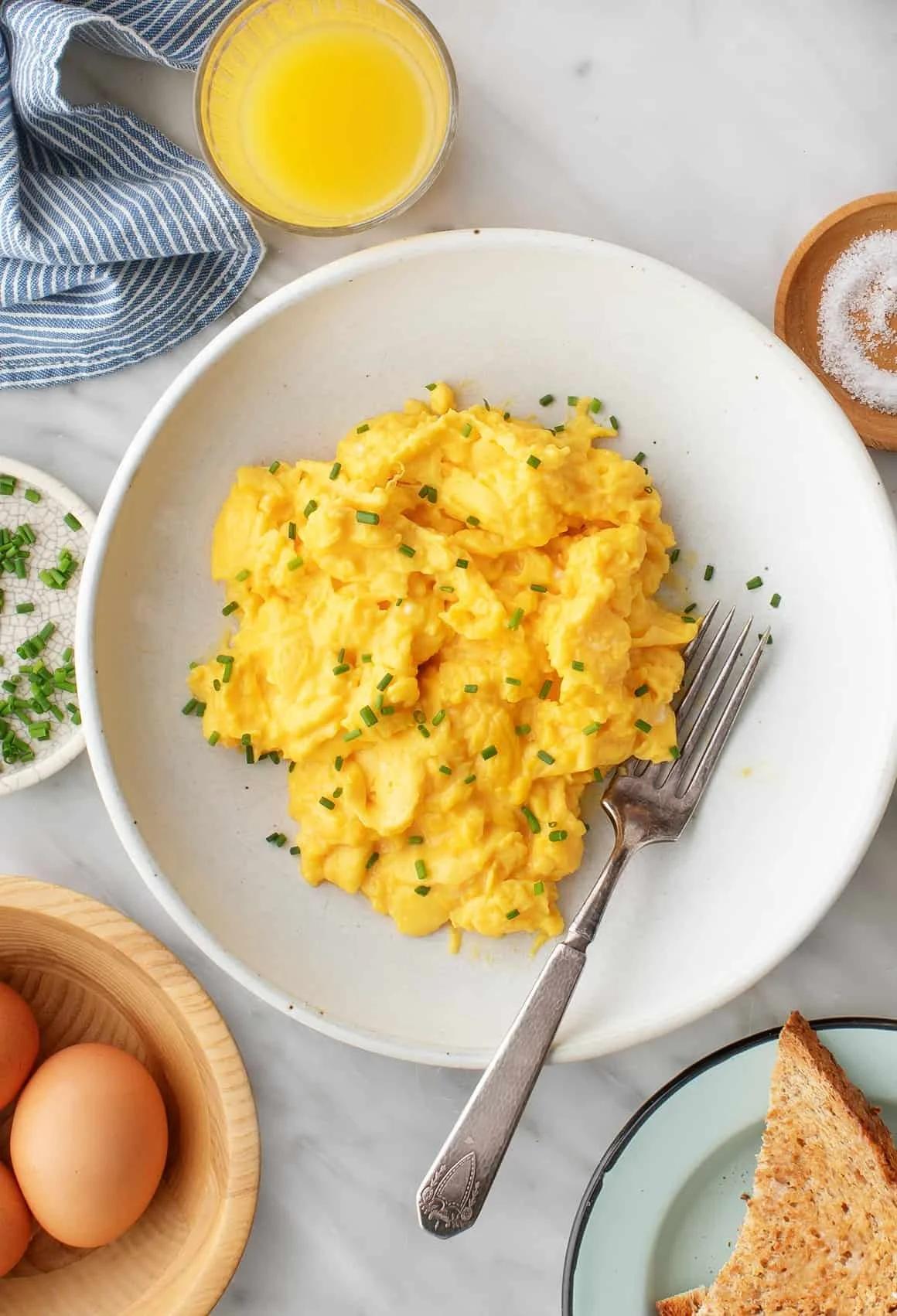 Scrambled eggs garnished with chopped chives on a white plate, with a fork resting on the plate, beside a glass of orange juice, a small bowl of salt, eggs in a wooden bowl, and toasted bread on a plate.