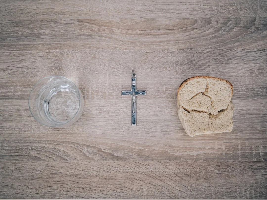 A clear glass of water, a metal cross pendant, and a piece of torn bread on a wooden surface.