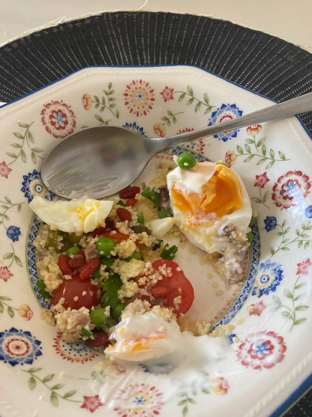 A decorative floral-patterned plate with remnants of a meal, including half a boiled egg, cherry tomatoes, green peas, rice, and some crumbs. A spoon rests on the plate.