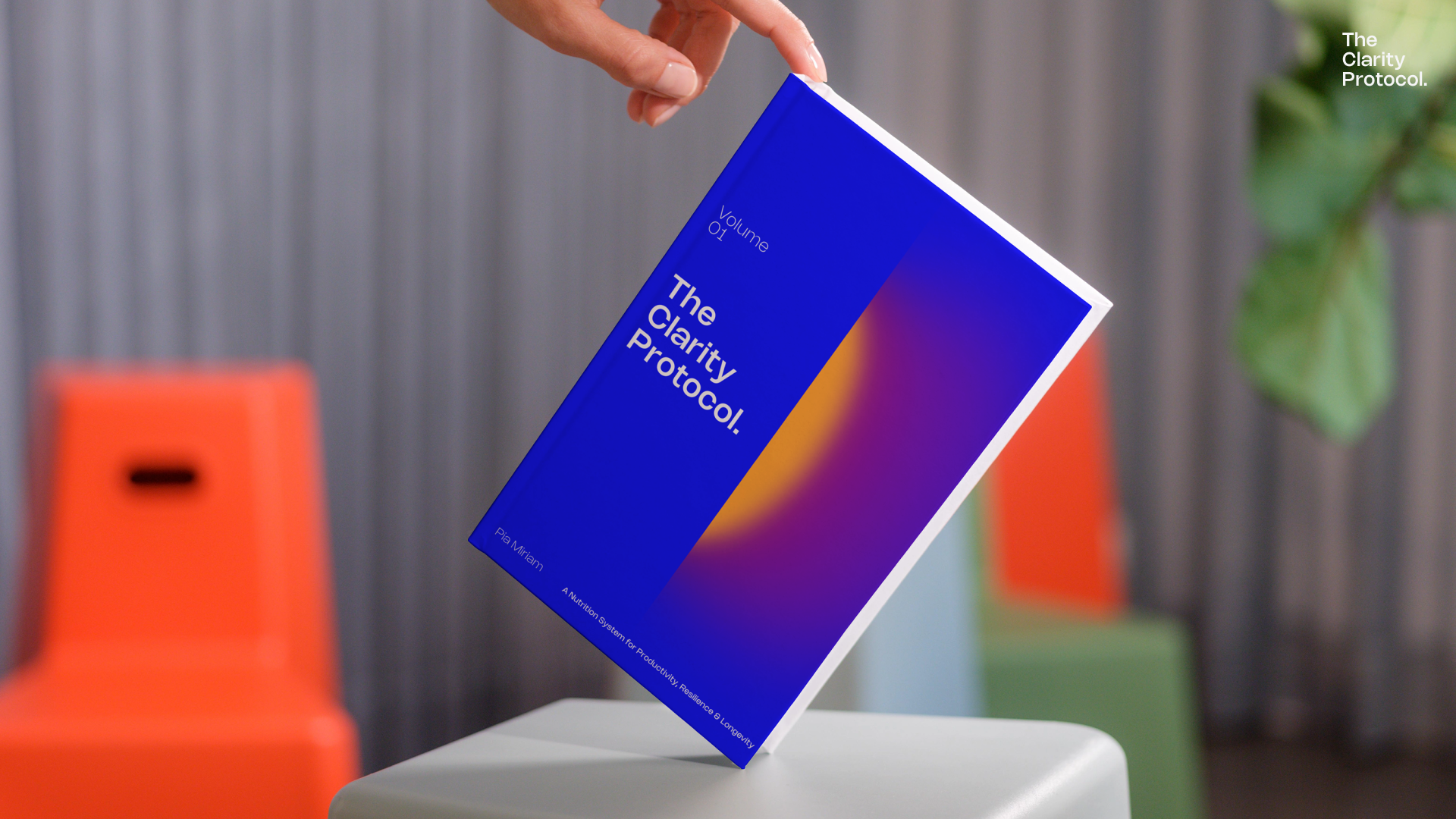 A book titled 'The Clarity Protocol' floating in mid-air. The cover is blue with white text and a colorful abstract design. In the background, there are orange, green, and gray chairs and a green plant.