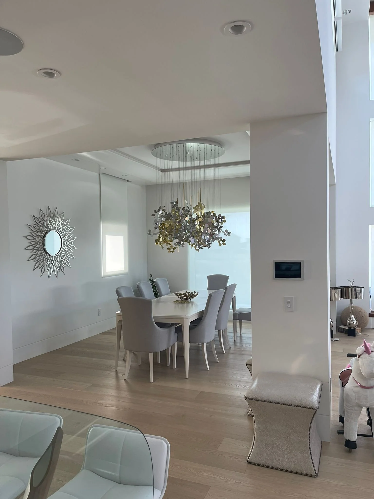 Elegant dining room with a white table surrounded by six white upholstered chairs, a modern chandelier with gold accents hanging overhead, a wall clock resembling a sunburst, and light wood flooring.