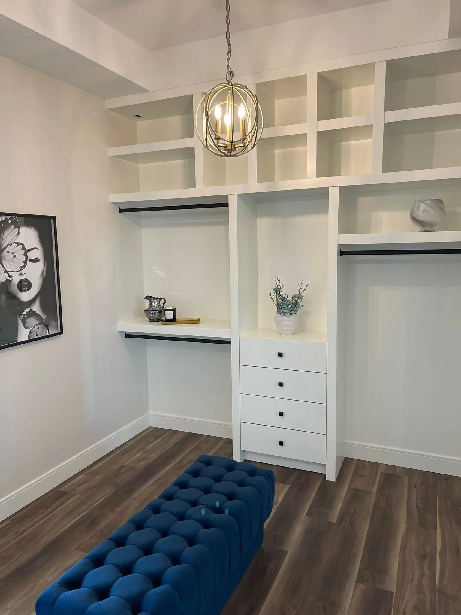 Interior view of a modern living space featuring a black and white framed art piece of a woman with butterfly wings on the left wall, white shelving with decorative items, a white chest of drawers with a potted succulent on top, a dark wooden floor, 