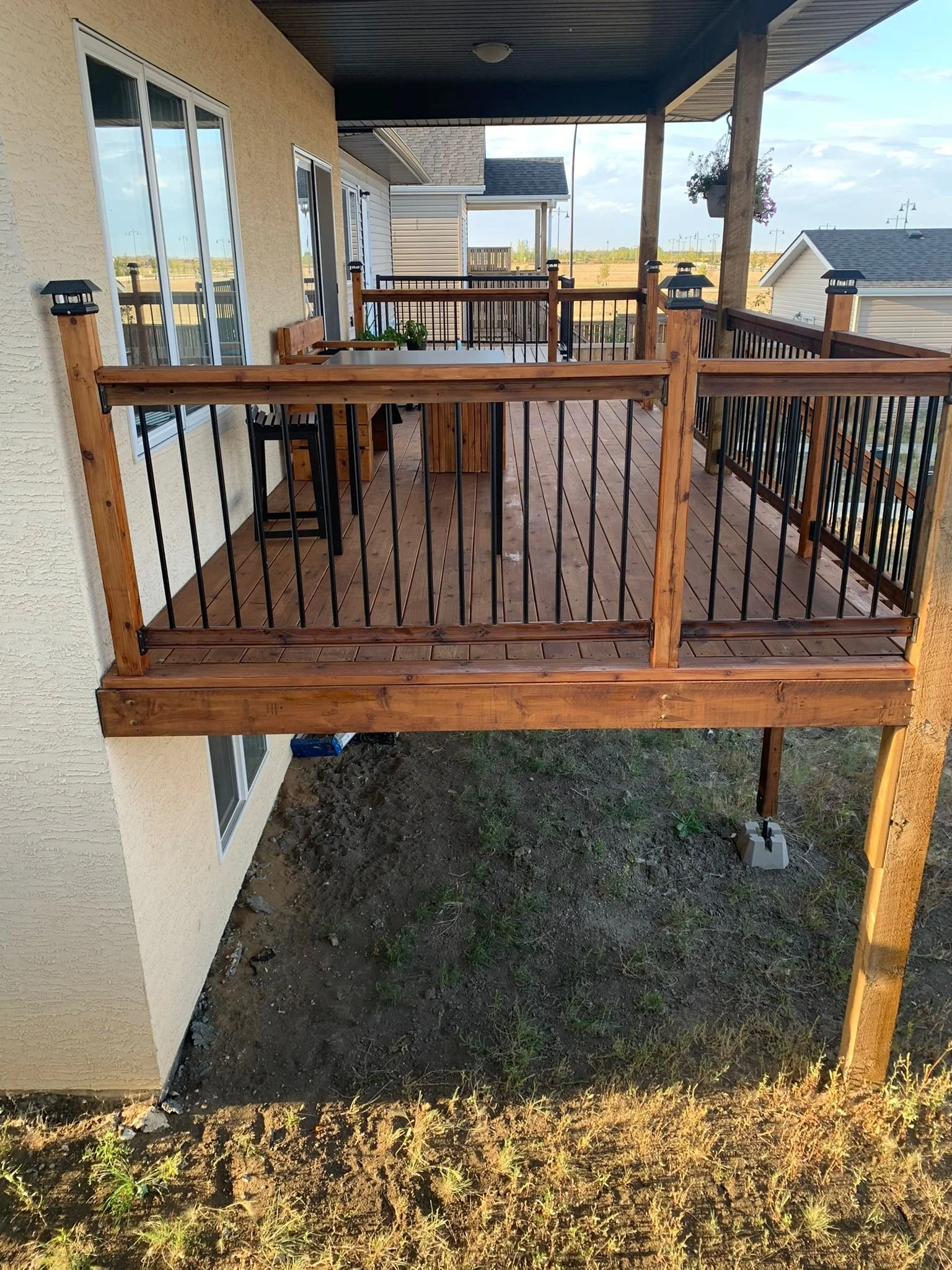 A wooden deck attached to a house with sliding glass doors, surrounded by a railing with black metal balusters and posts topped with black lantern-style lights. The deck overlooks a dirt ground with sparse grass and a distant view of a residential ne