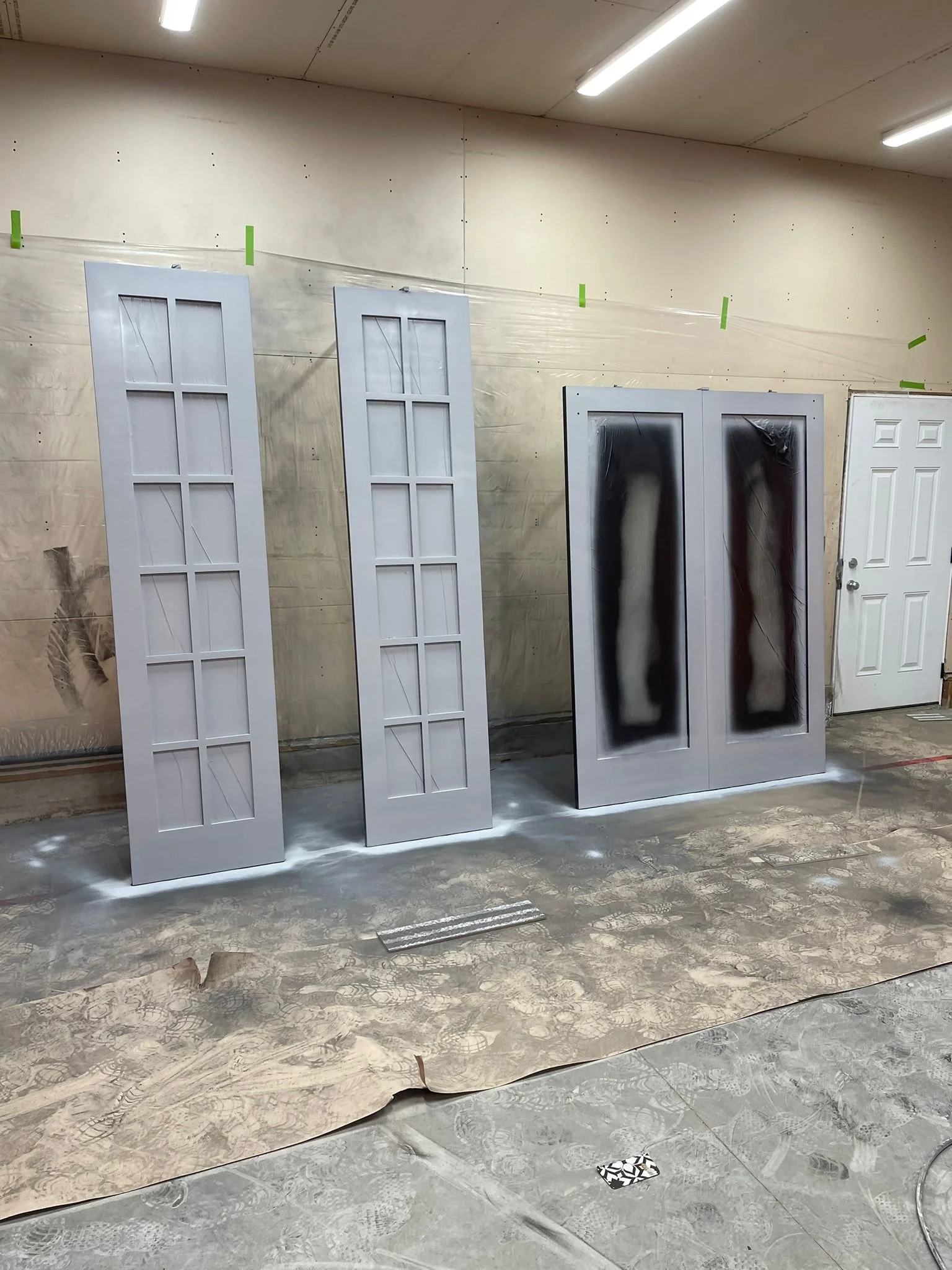Four doors leaning against a wall in a workshop, with a large sheet of patterned material on the floor and ceiling lights overhead.