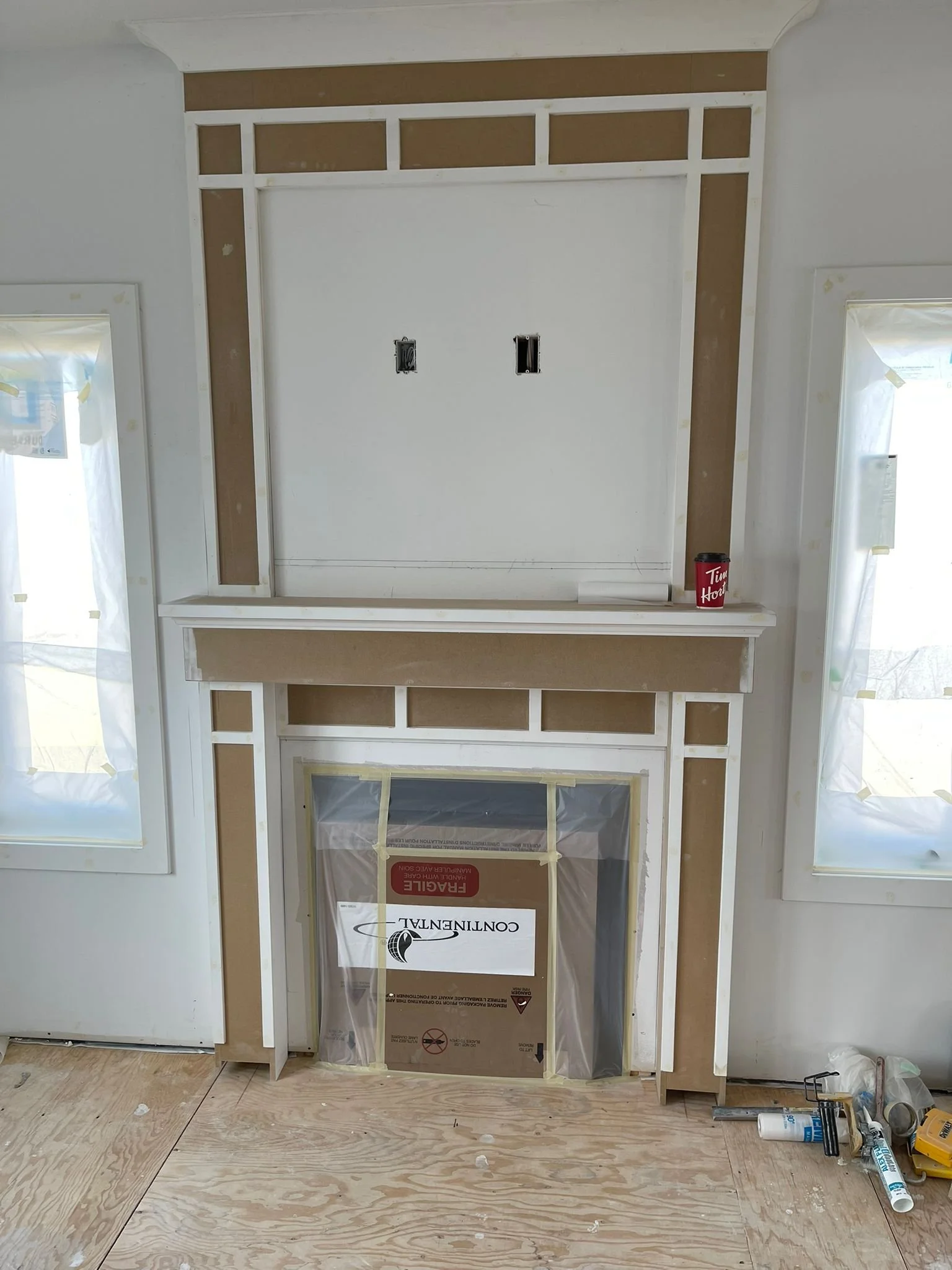 Room with construction in progress, featuring a wall with framing, electrical outlets, and a cardboard box with plastic covering over a doorway, surrounded by construction tools and materials.